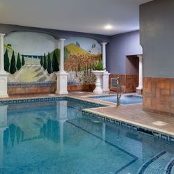 Indoor swimming pool with mosaic tiles, a hot tub area, and decorative mural on the wall featuring trees, columns, and landscape art.