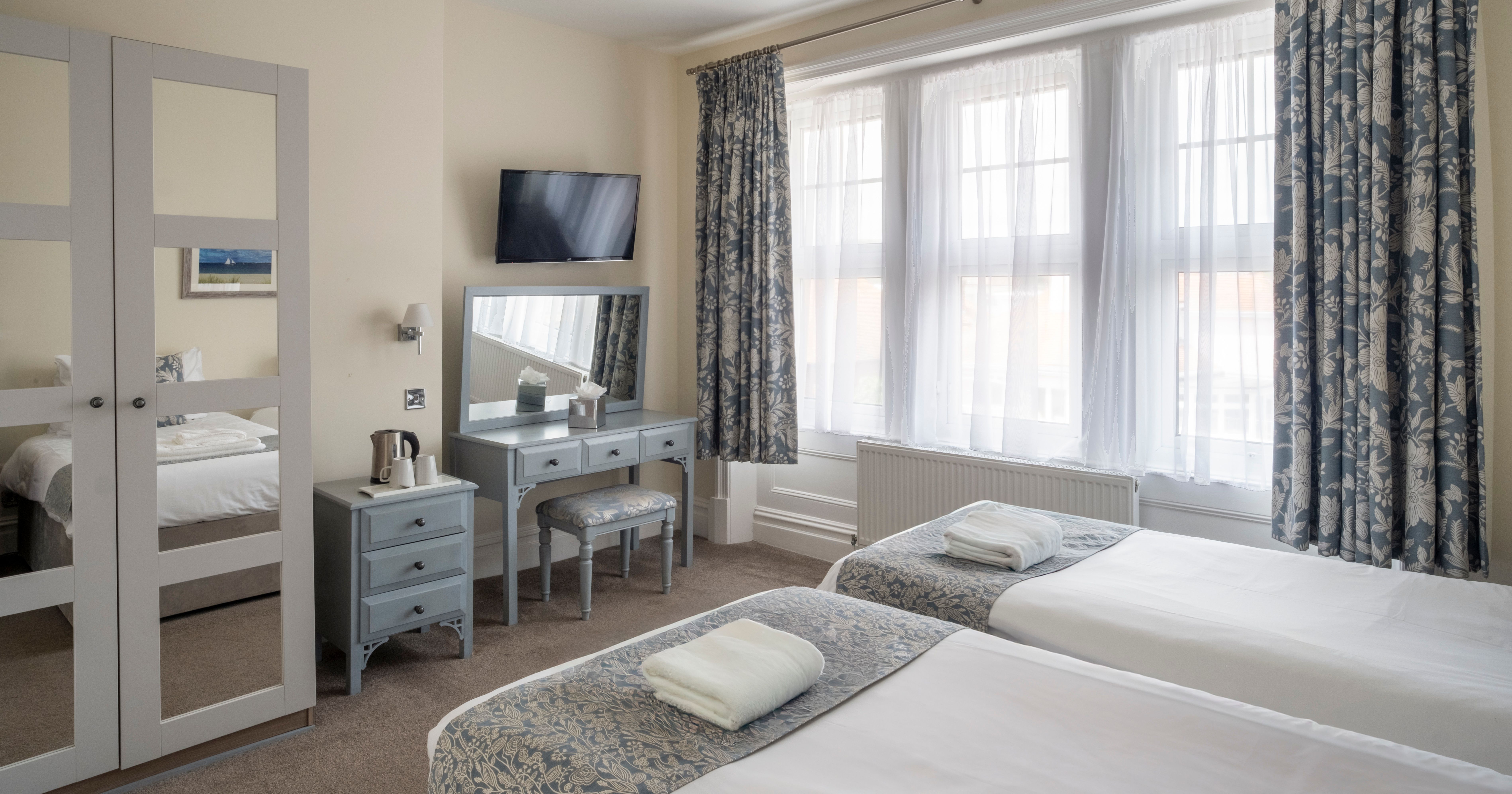 Bright hotel room with two single beds, dressing table, TV, wardrobe, and large windows with floral curtains