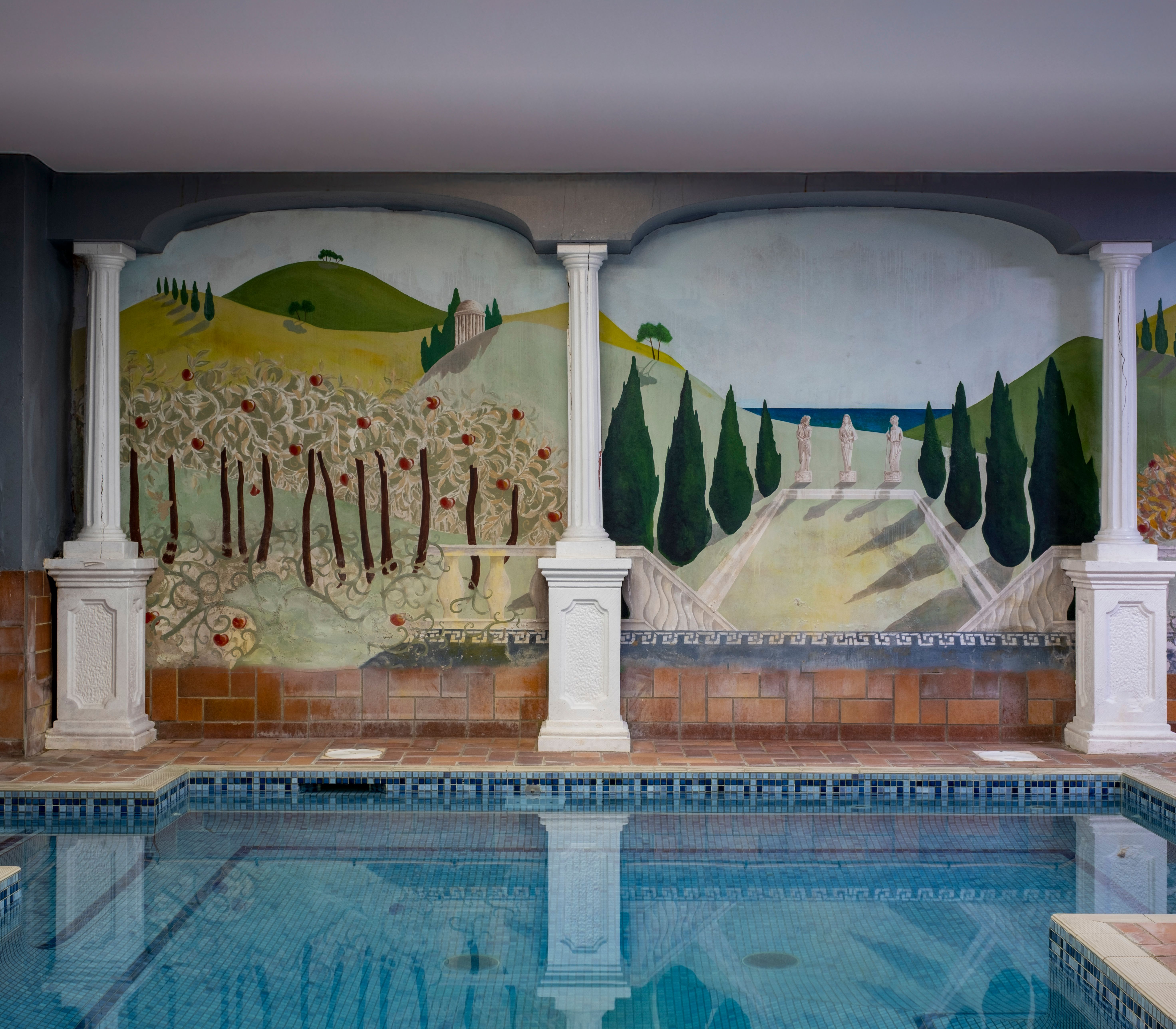 Indoor swimming pool with a decorative wall mural featuring columns, trees, and a painted landscape of hills, cypress trees, and statues.
