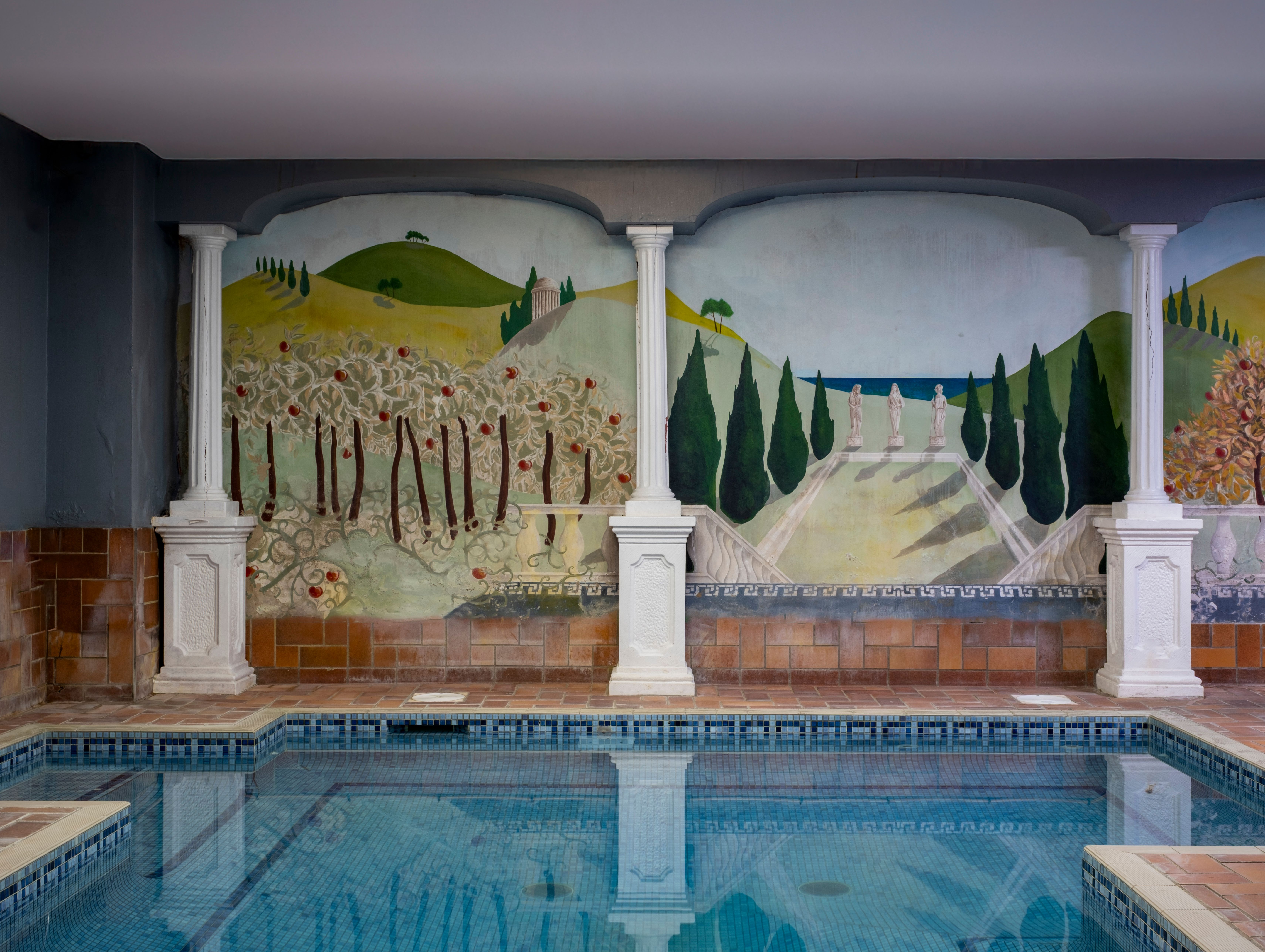 Indoor swimming pool with a decorative wall mural featuring columns, trees, and a painted landscape of hills, cypress trees, and statues.