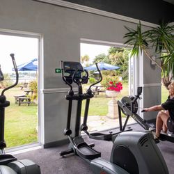 A woman using a rowing machine in a gym with large open doors leading to an outdoor garden area with benches and umbrellas.