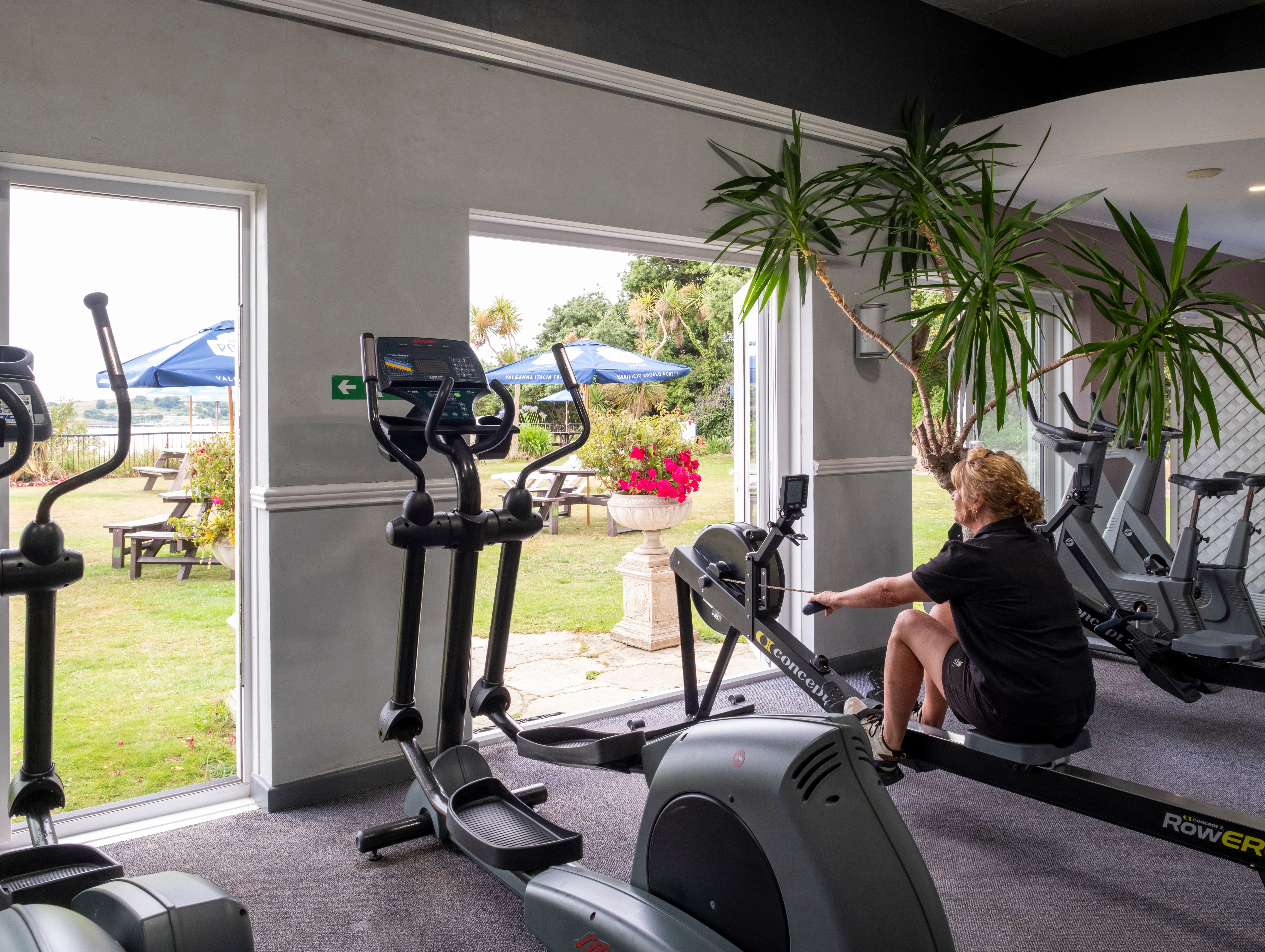 A woman using a rowing machine in a gym with large open doors leading to an outdoor garden area with benches and umbrellas.