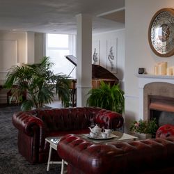 Elegant lounge area with red leather sofas, a coffee table set for tea, a fireplace with candles, a large decorative wall clock, green plants, and a grand piano near a window.