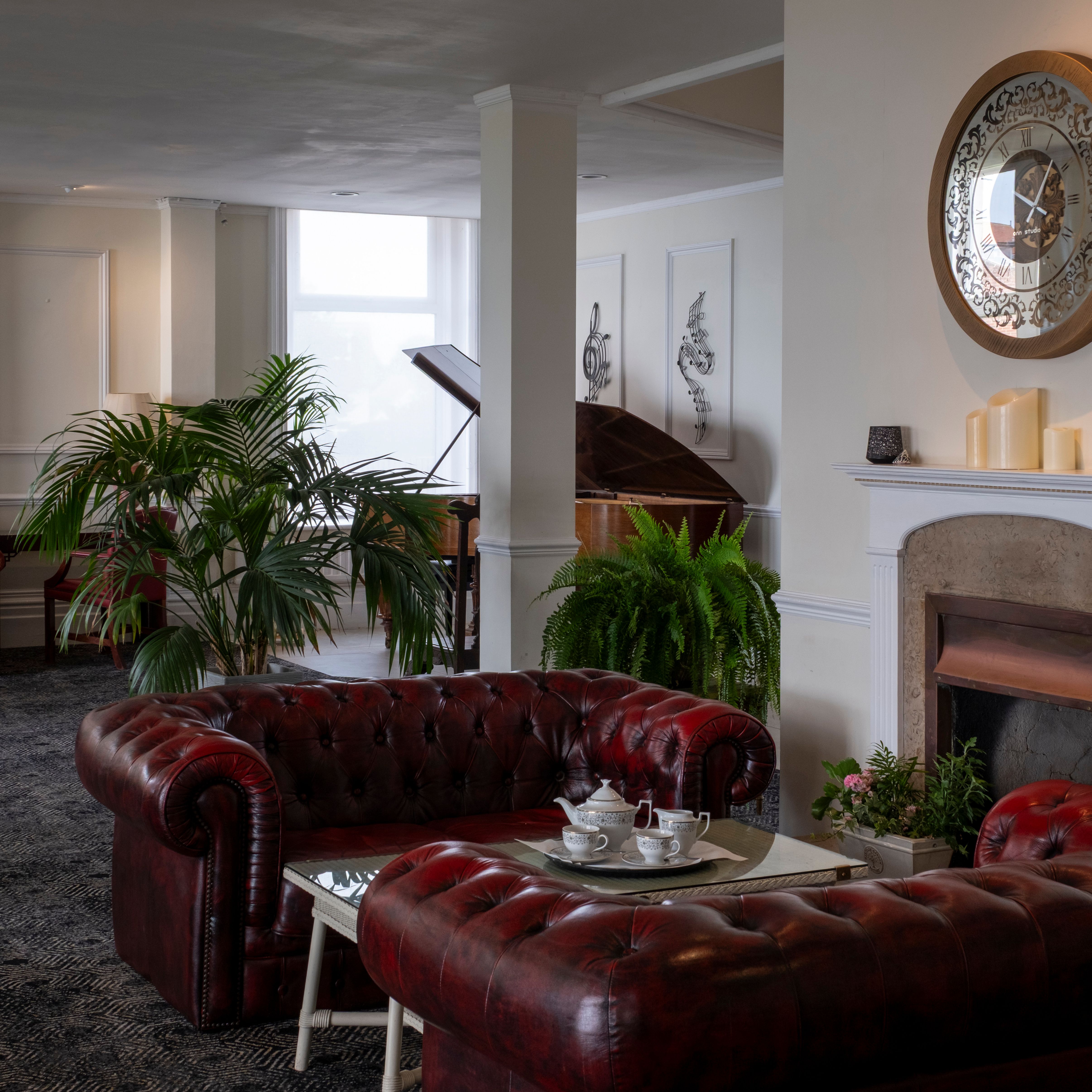 Elegant lounge area with red leather sofas, a coffee table set for tea, a fireplace with candles, a large decorative wall clock, green plants, and a grand piano near a window.