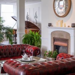 Elegant living room with red leather sofas, a glass coffee table set for tea, a fireplace with a large decorative mirror above, green plants, and a grand piano in the background.