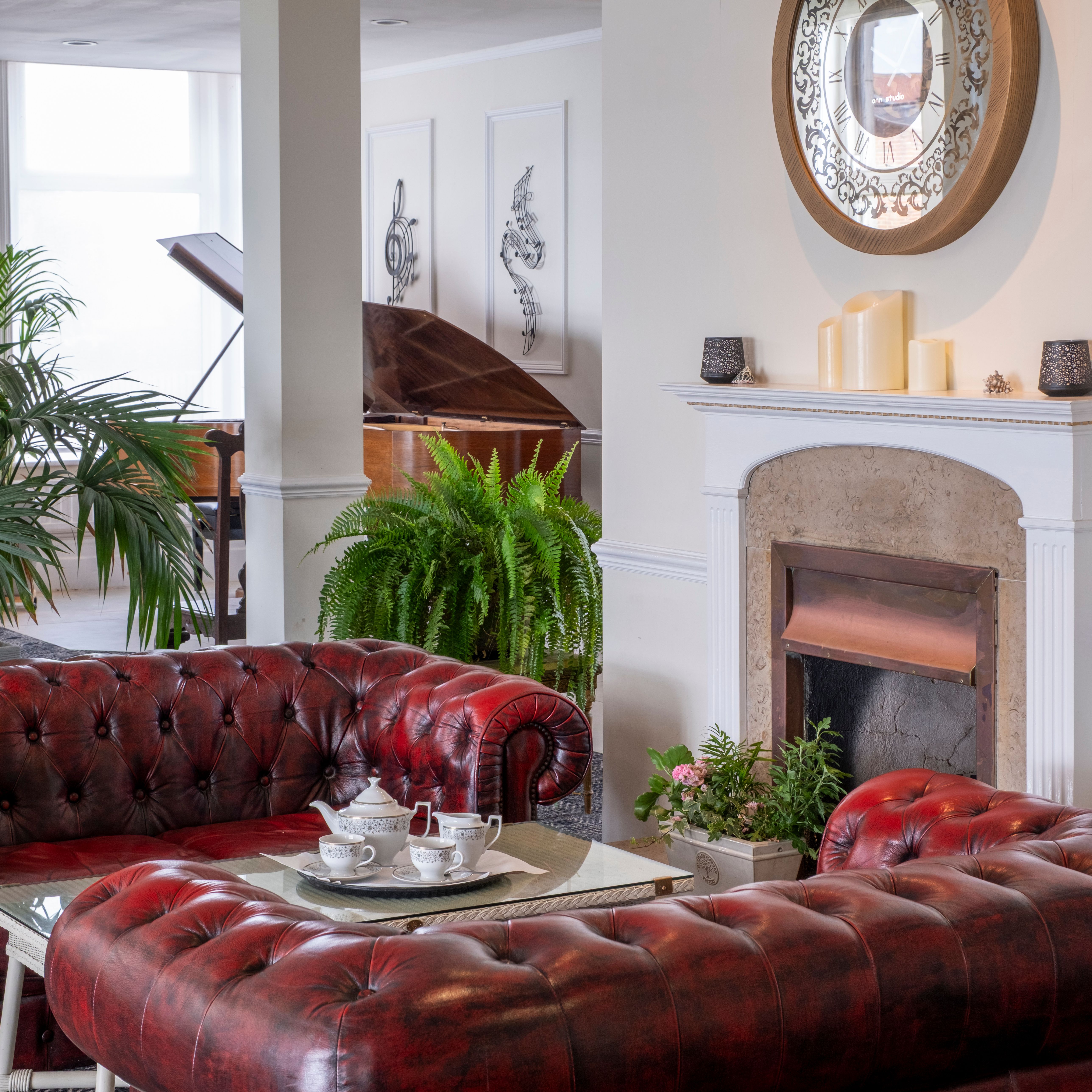 Elegant living room with red leather sofas, a glass coffee table set for tea, a fireplace with a large decorative mirror above, green plants, and a grand piano in the background.