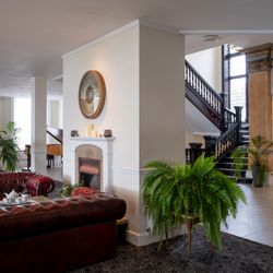 Elegant hotel lobby with red leather sofas, a fireplace, large decorative clock, indoor plants, staircase with dark wood banister, grand piano, and ornate gold mirror.