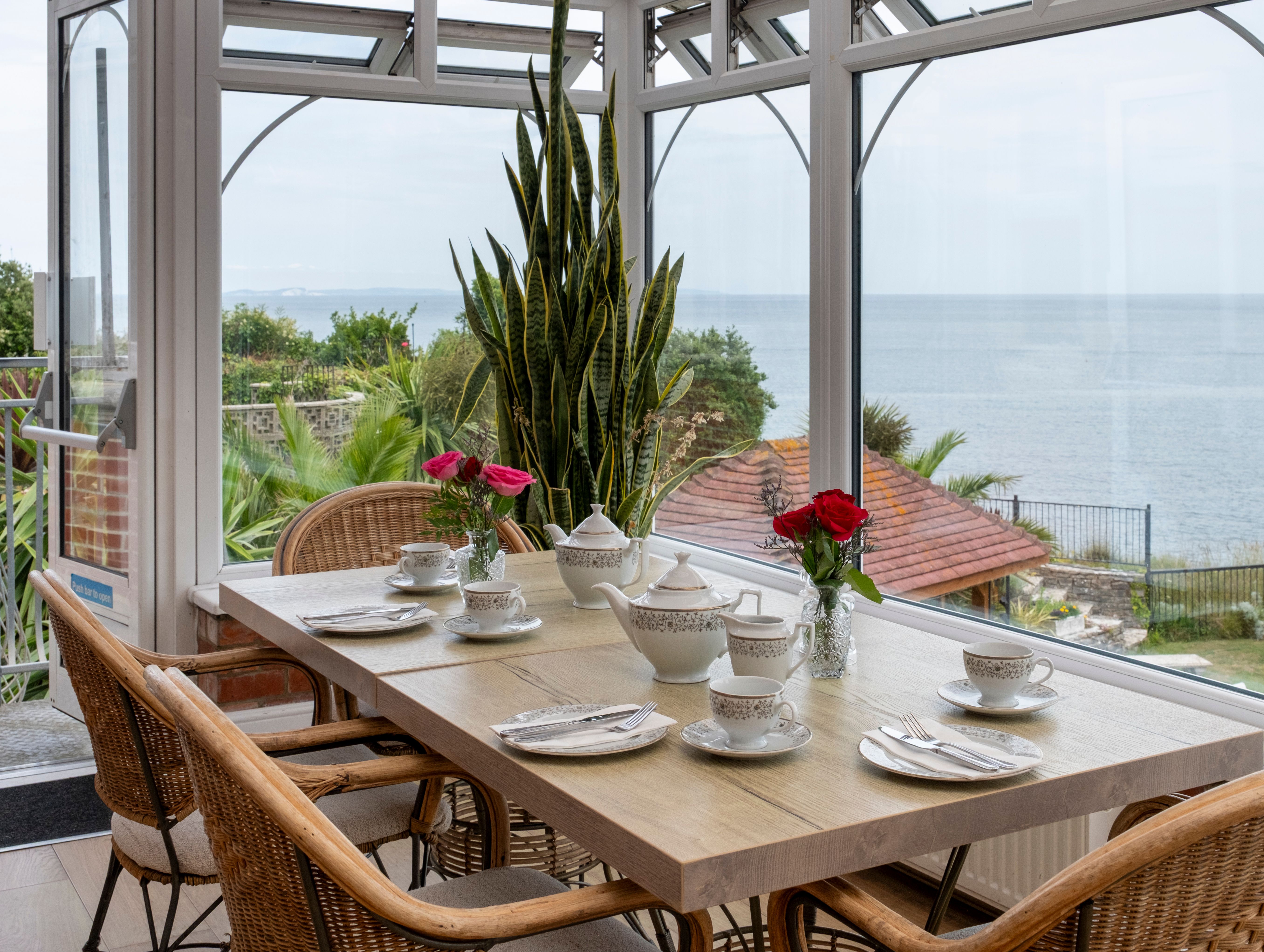 Elegant table set for tea with china cups, teapots, and vases of red roses in a sunroom overlooking a scenic ocean view.