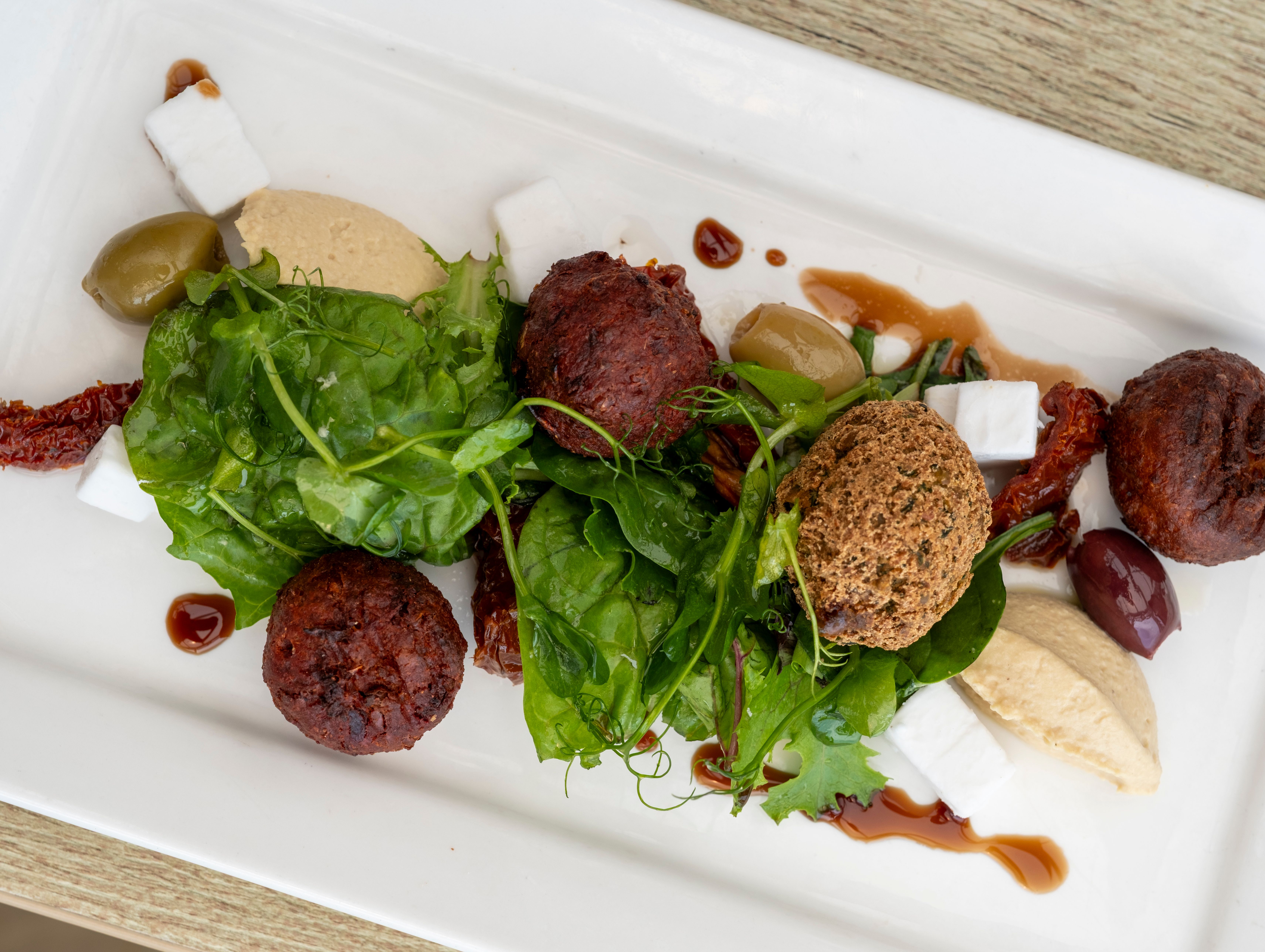 A plated Mediterranean dish featuring falafel balls, mixed greens, hummus, feta cheese cubes, olives, sun-dried tomatoes, and a drizzle of sauce on a white rectangular plate.