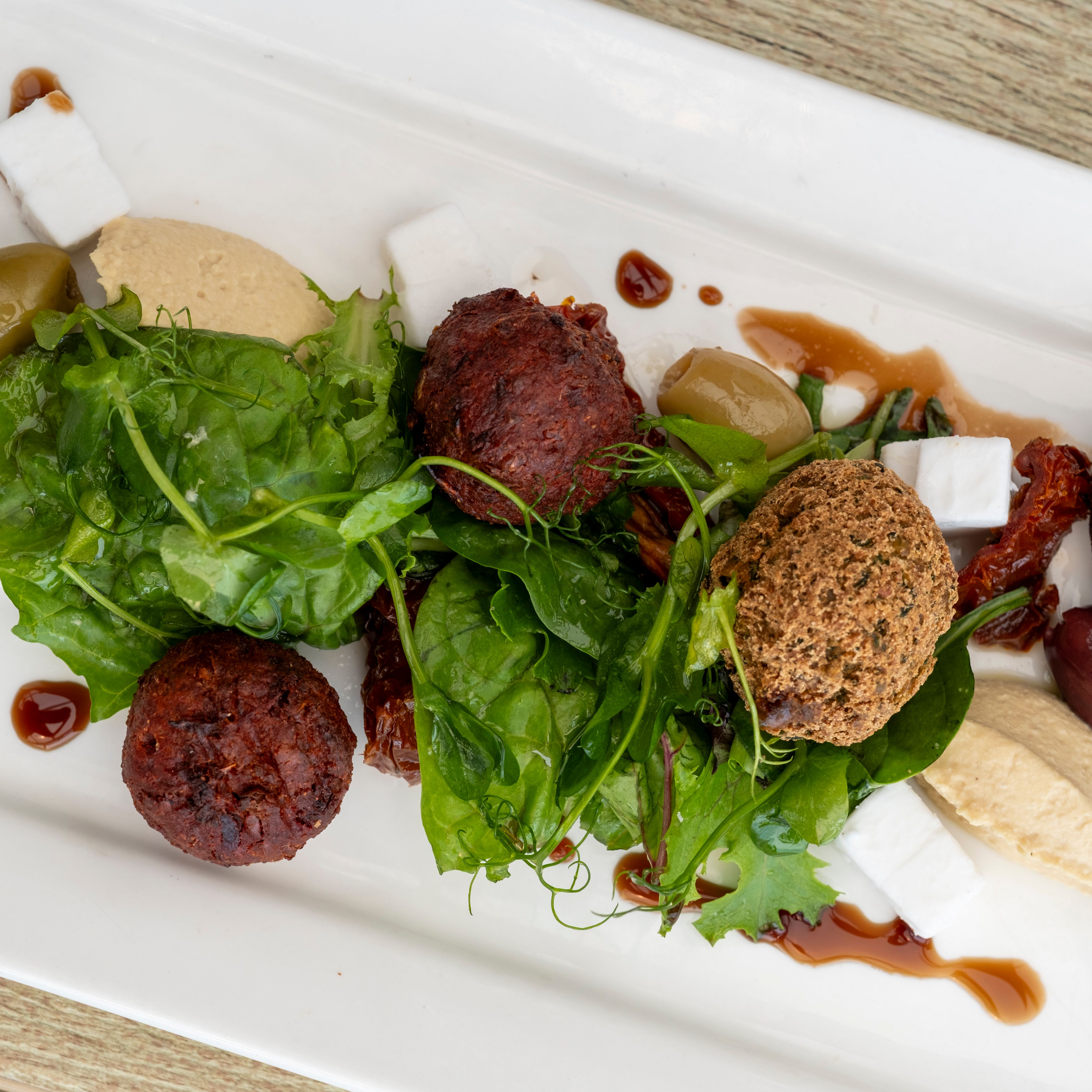 A plated Mediterranean dish featuring falafel balls, mixed greens, hummus, feta cheese cubes, olives, sun-dried tomatoes, and a drizzle of sauce on a white rectangular plate.