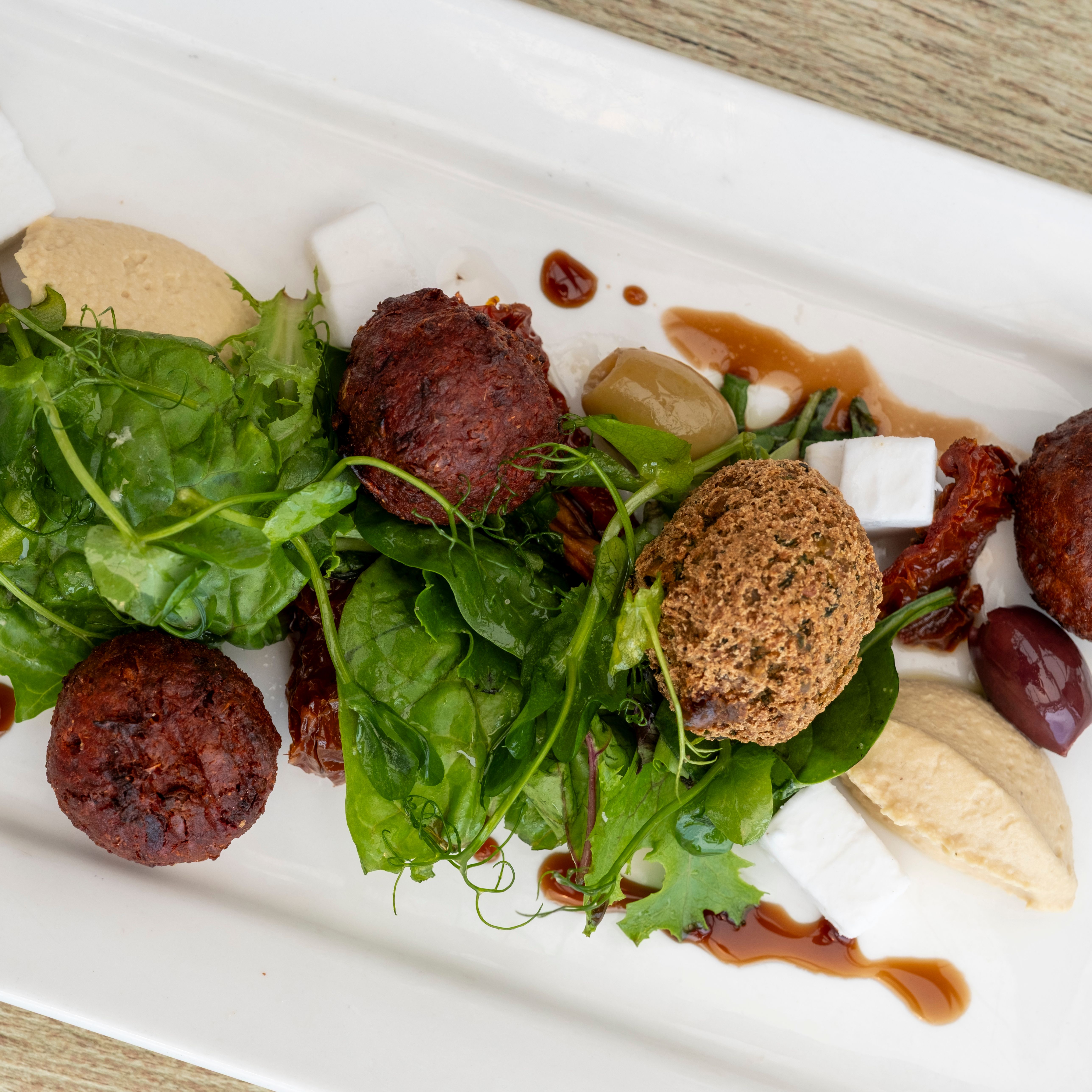 A plated Mediterranean dish featuring falafel balls, mixed greens, hummus, feta cheese cubes, olives, sun-dried tomatoes, and a drizzle of sauce on a white rectangular plate.