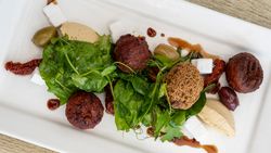 A plated Mediterranean dish featuring falafel balls, mixed greens, hummus, feta cheese cubes, olives, sun-dried tomatoes, and a drizzle of sauce on a white rectangular plate.