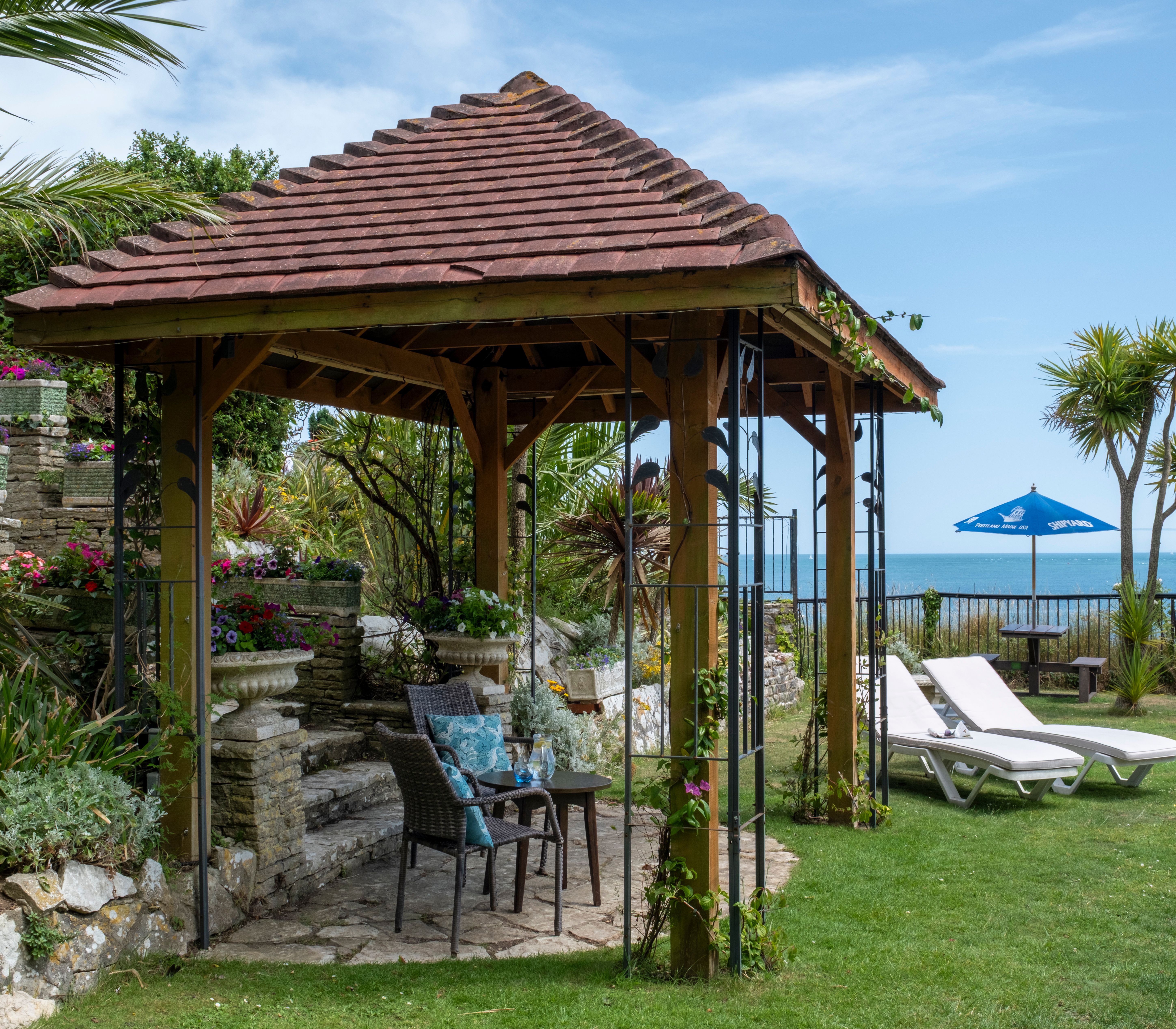 A wooden gazebo in a garden with lounge chairs, stone steps, potted flowers, palm trees, and a view of the sea with a blue sky.