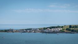 Coastal village with houses and a pier by the sea, green hills in the background, and several boats on the water under a blue sky.