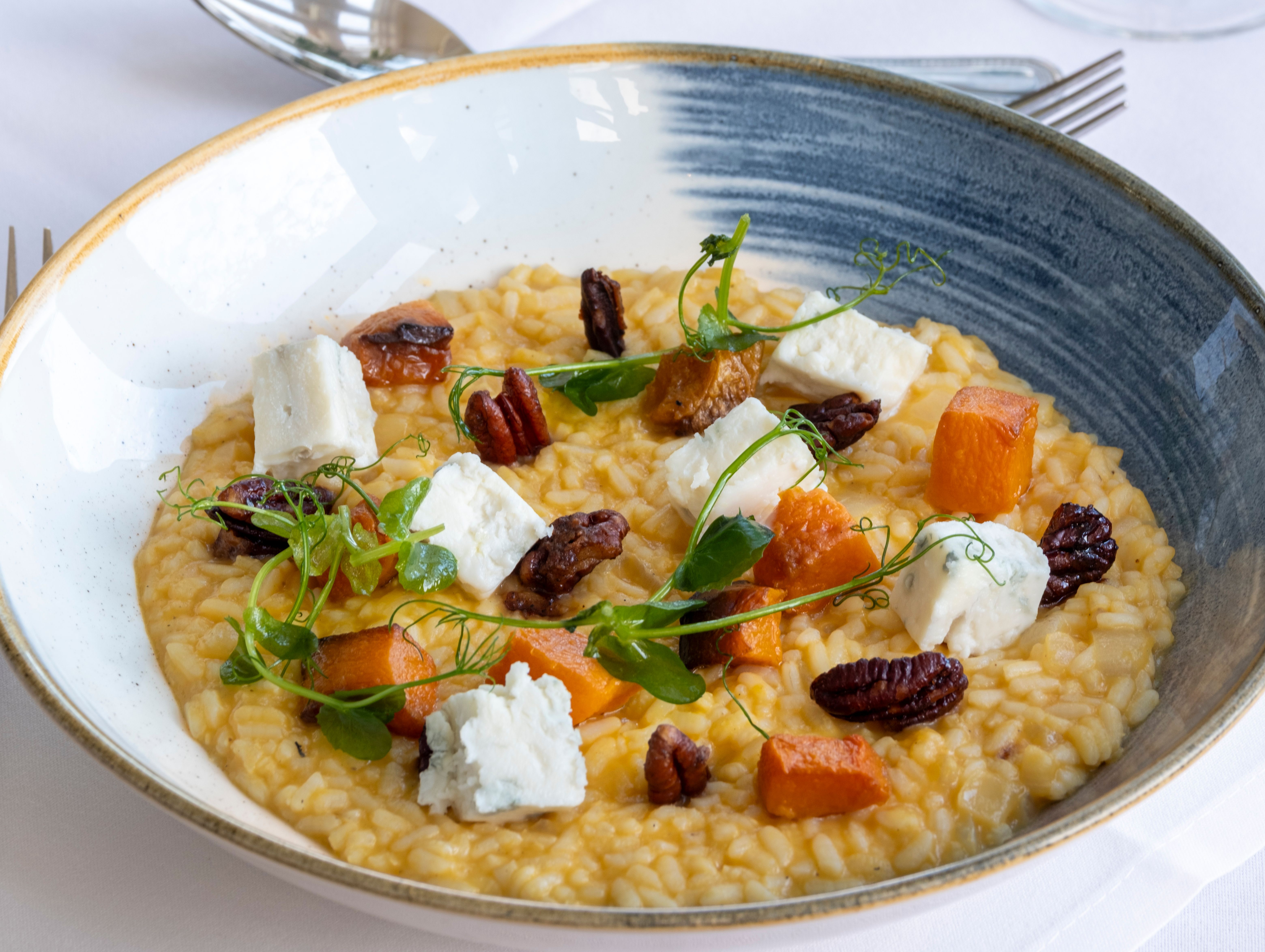 A plate of creamy risotto garnished with roasted butternut squash, pecans, cubes of cheese, and microgreens.