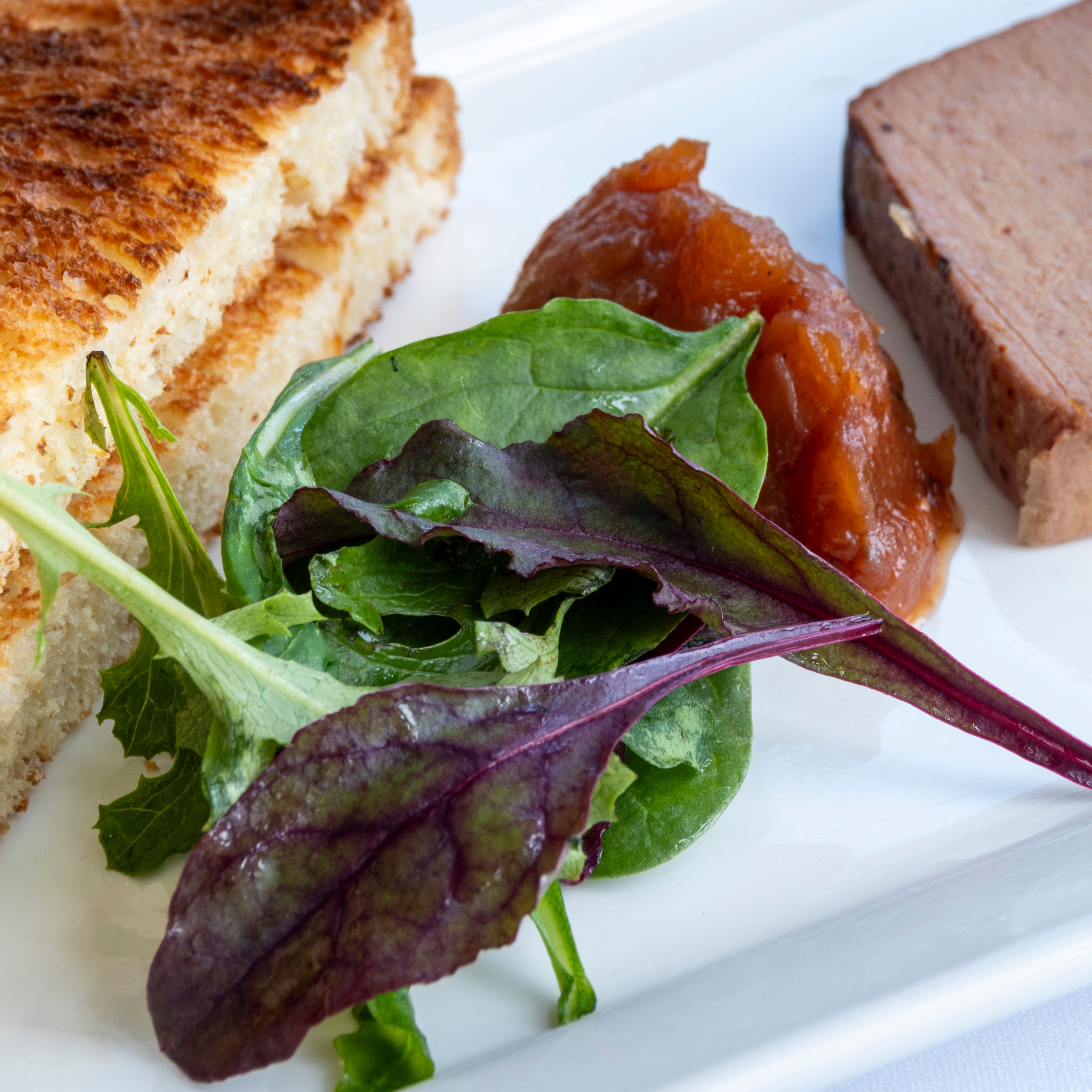 A plate with toasted bread slices, mixed leafy greens, a dollop of chutney, and a slice of pâté.