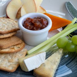 A plate with assorted crackers, cheese wedges, green grapes, celery sticks, apple slices, a small dish of chutney or relish, and a knife.