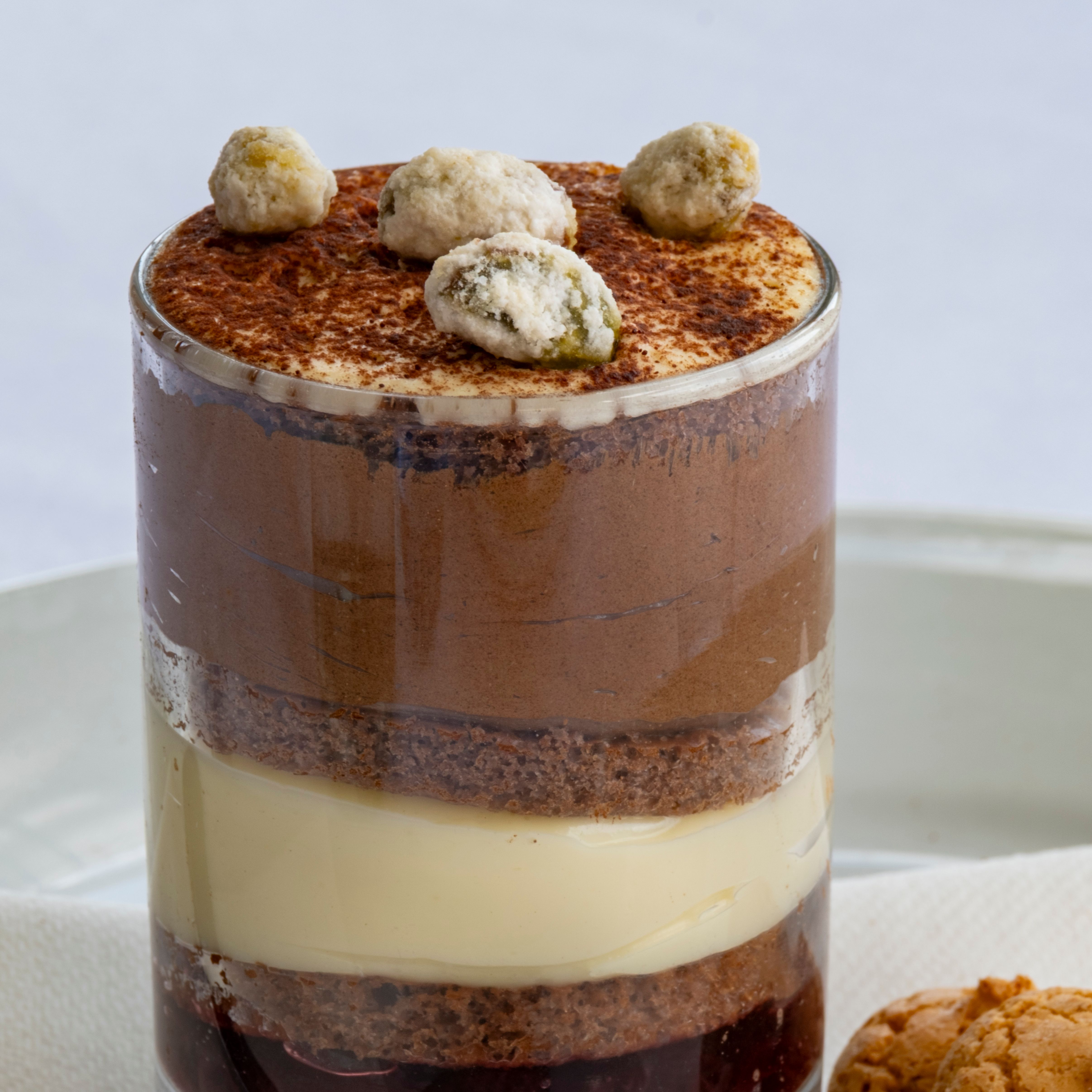 Layered dessert in a glass with chocolate, cream, cake, and berry layers, topped with cookies. A spoon and two cookies are beside it on a white napkin.