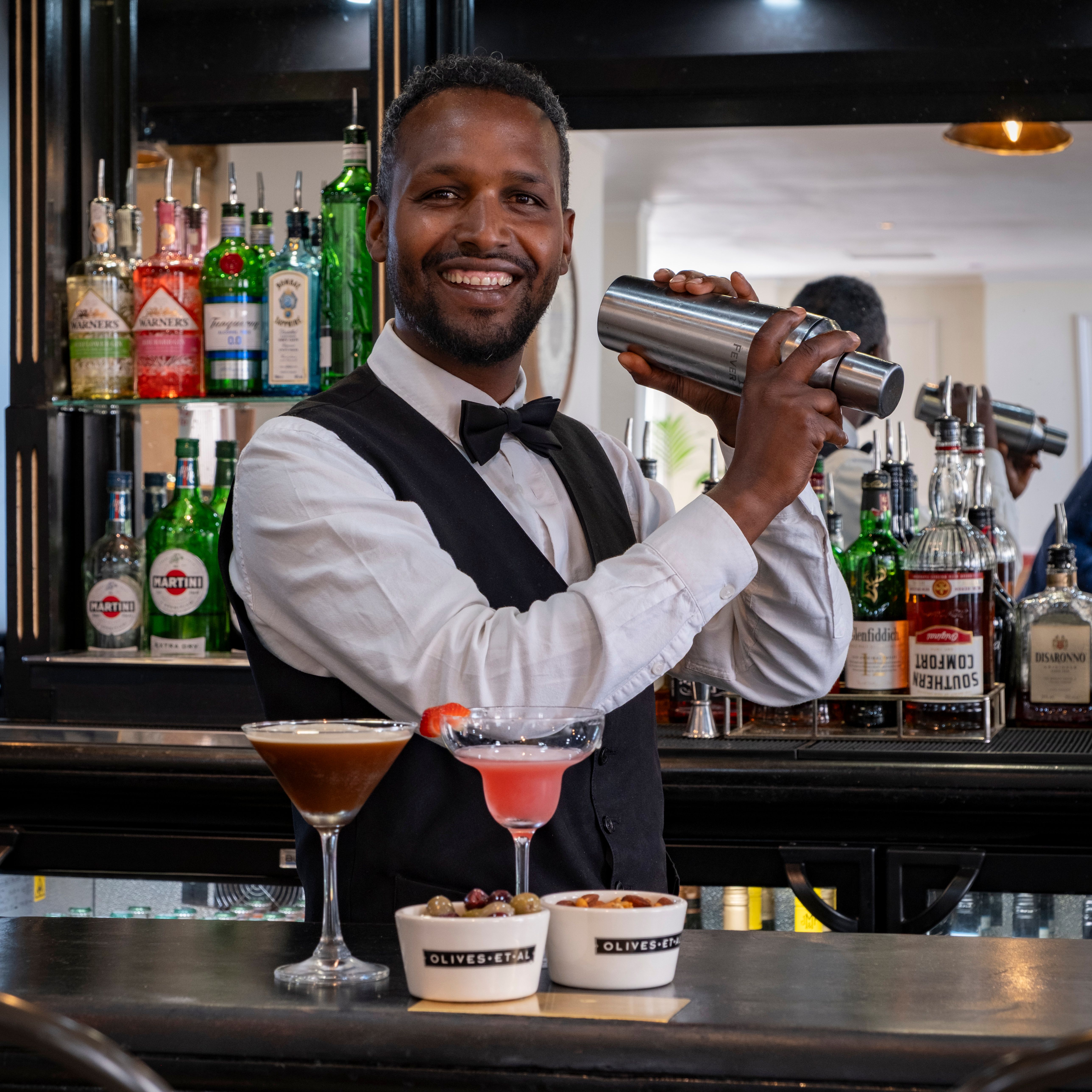Smiling bartender shaking a cocktail shaker behind a bar, with various bottles of alcohol on the shelves and two cocktails and bowls of snacks on the counter.