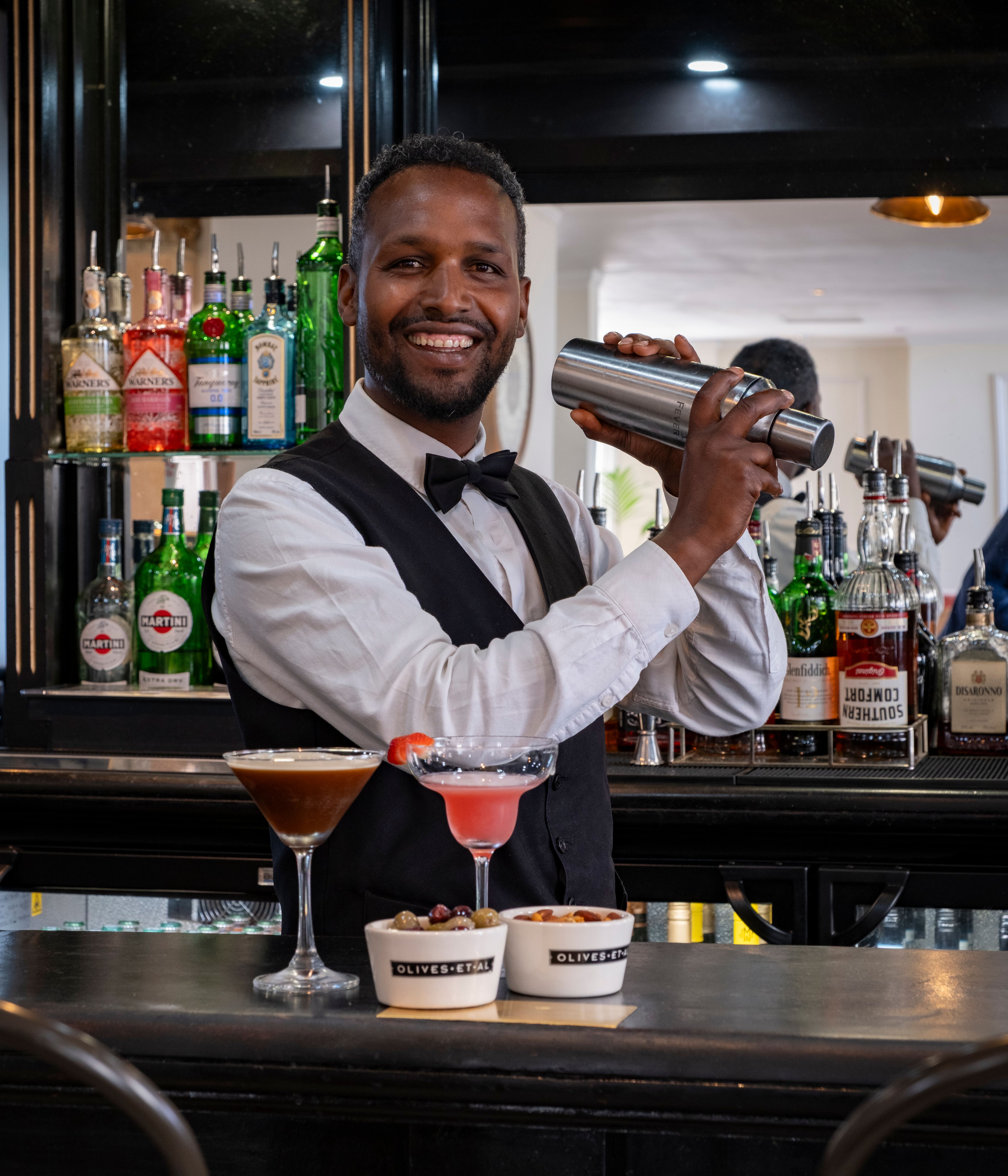 Smiling bartender shaking a cocktail shaker behind a bar, with various bottles of alcohol on the shelves and two cocktails and bowls of snacks on the counter.