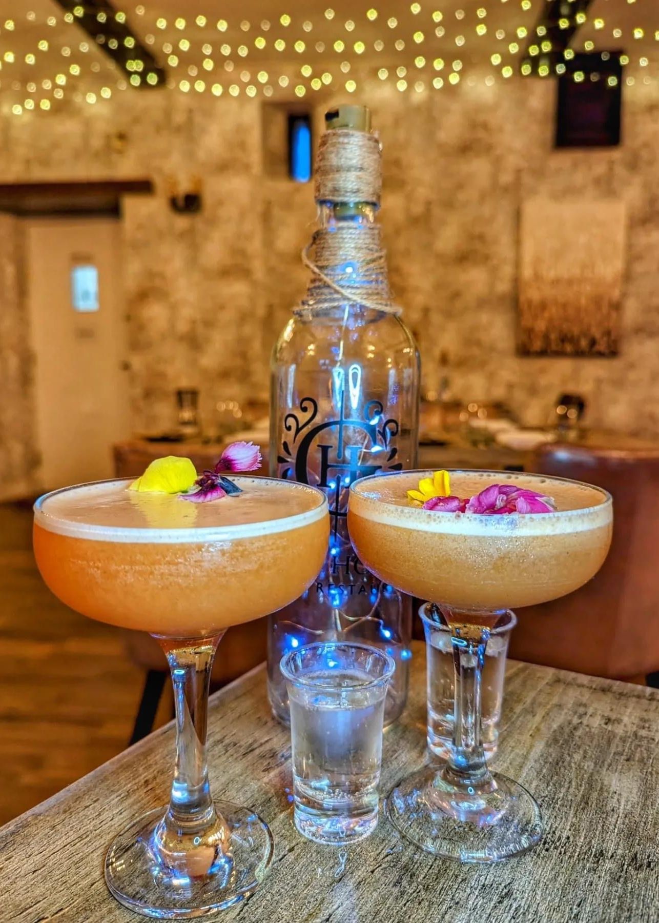 Two elegant cocktails garnished with edible flowers, a shot glass, and a decorative bottle on a rustic table with fairy lights in the background.
