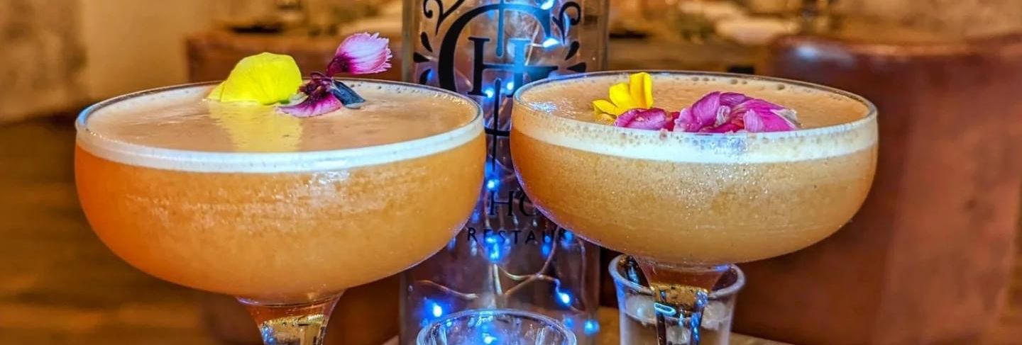 Two elegant cocktails garnished with edible flowers, a shot glass, and a decorative bottle on a rustic table with fairy lights in the background.