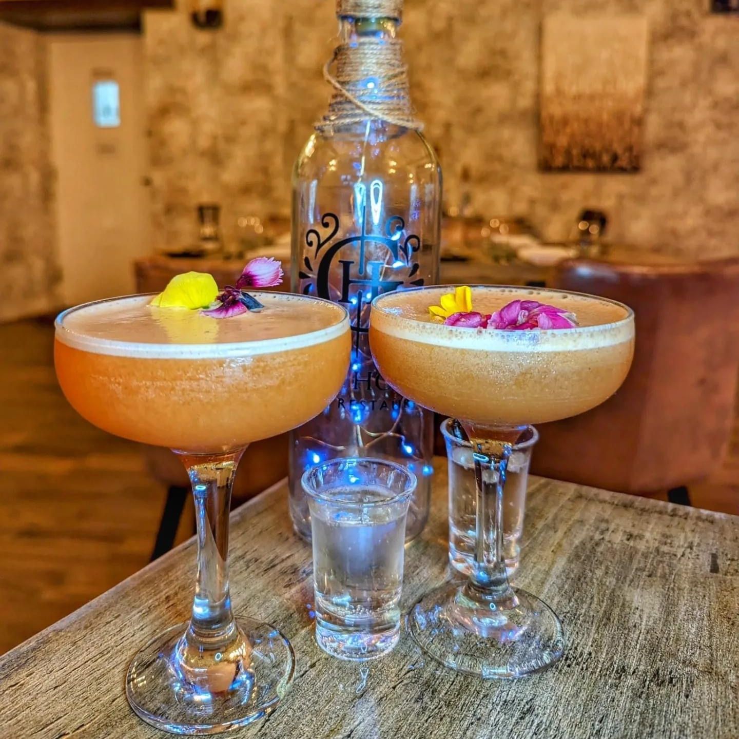 Two elegant cocktails garnished with edible flowers, a shot glass, and a decorative bottle on a rustic table with fairy lights in the background.