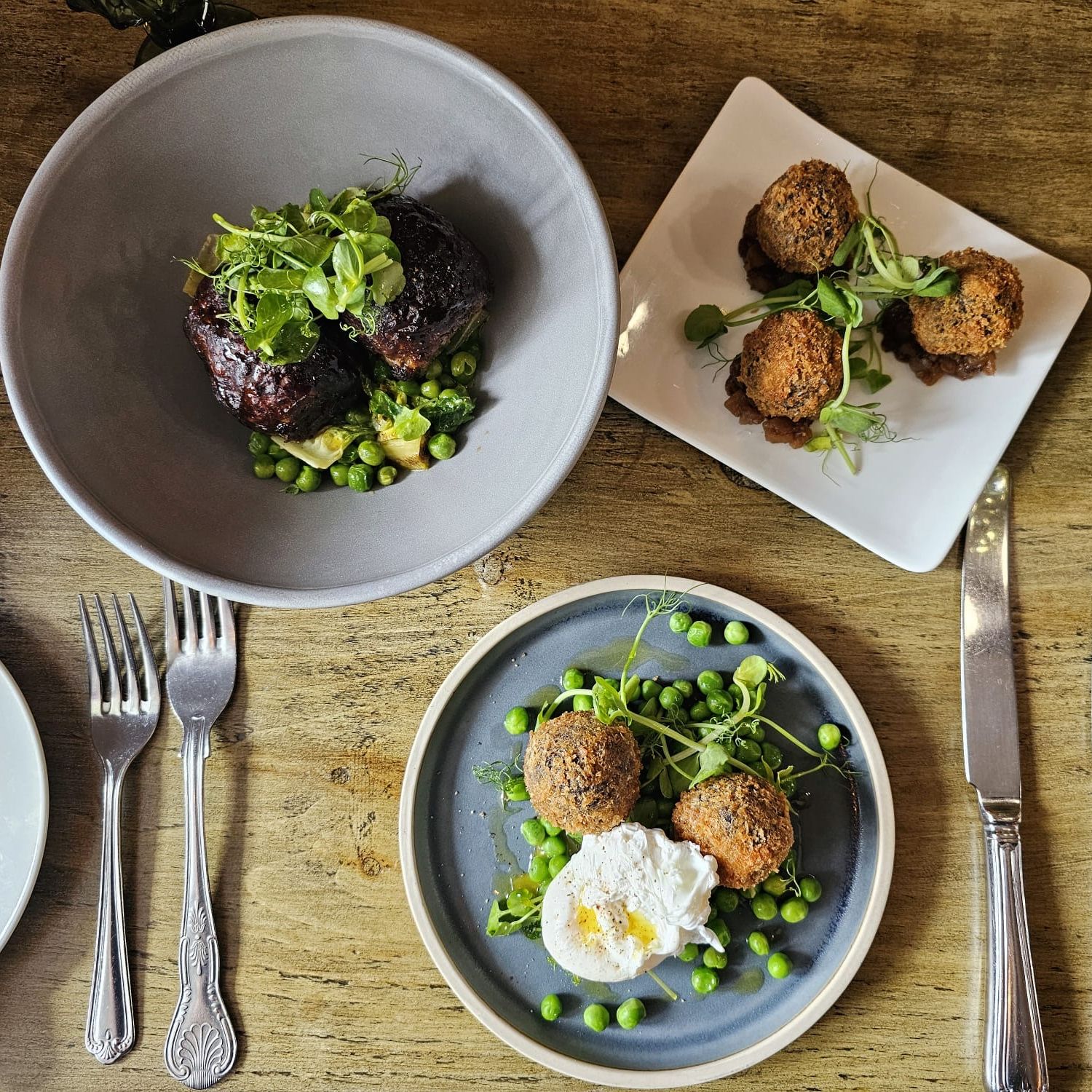 A rustic table setting with three plates of gourmet food including croquettes, meatballs, peas, and a poached egg.