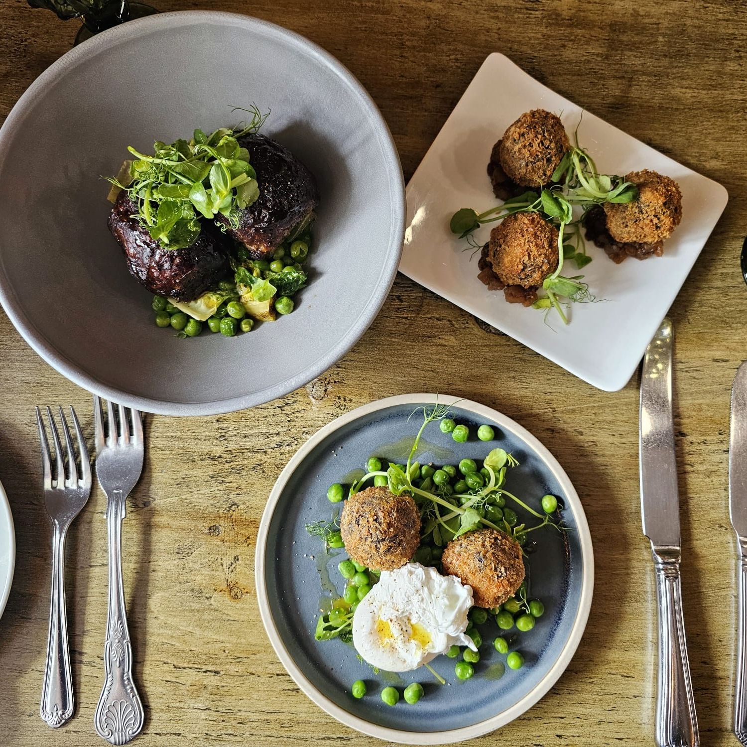 A rustic table setting with three plates of gourmet food including croquettes, meatballs, peas, and a poached egg.