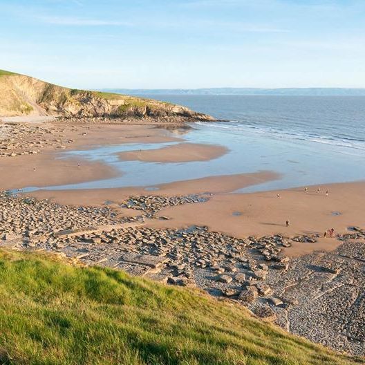 A scenic coastal view with sandy beach, rocky shore, and grassy cliffs under a clear blue sky