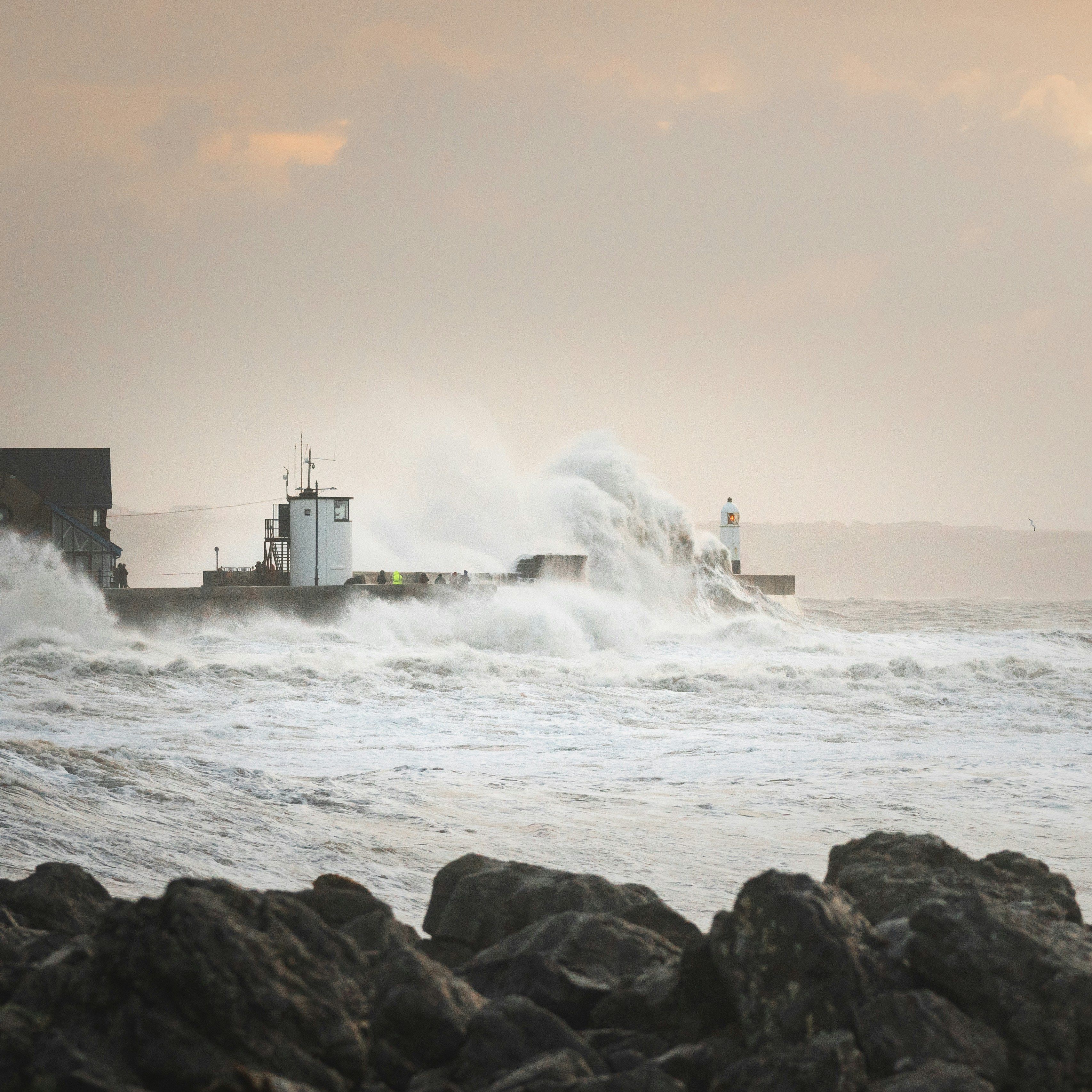 Large crashing waves against a pier with a lighthouse during a stormy sunset.