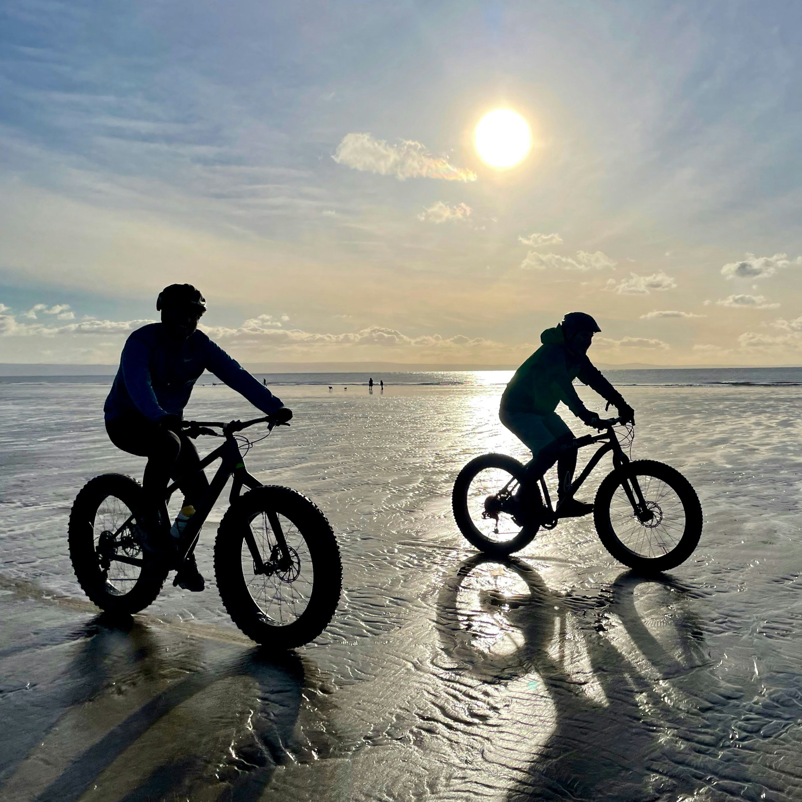 Two people riding fat tire bikes on a wet sandy beach during sunset with the sun glowing and casting long shadows.