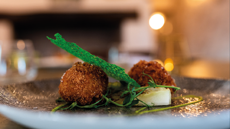 Gourmet plated dish featuring two breaded and fried balls garnished with fresh greens and a crisp green decoration