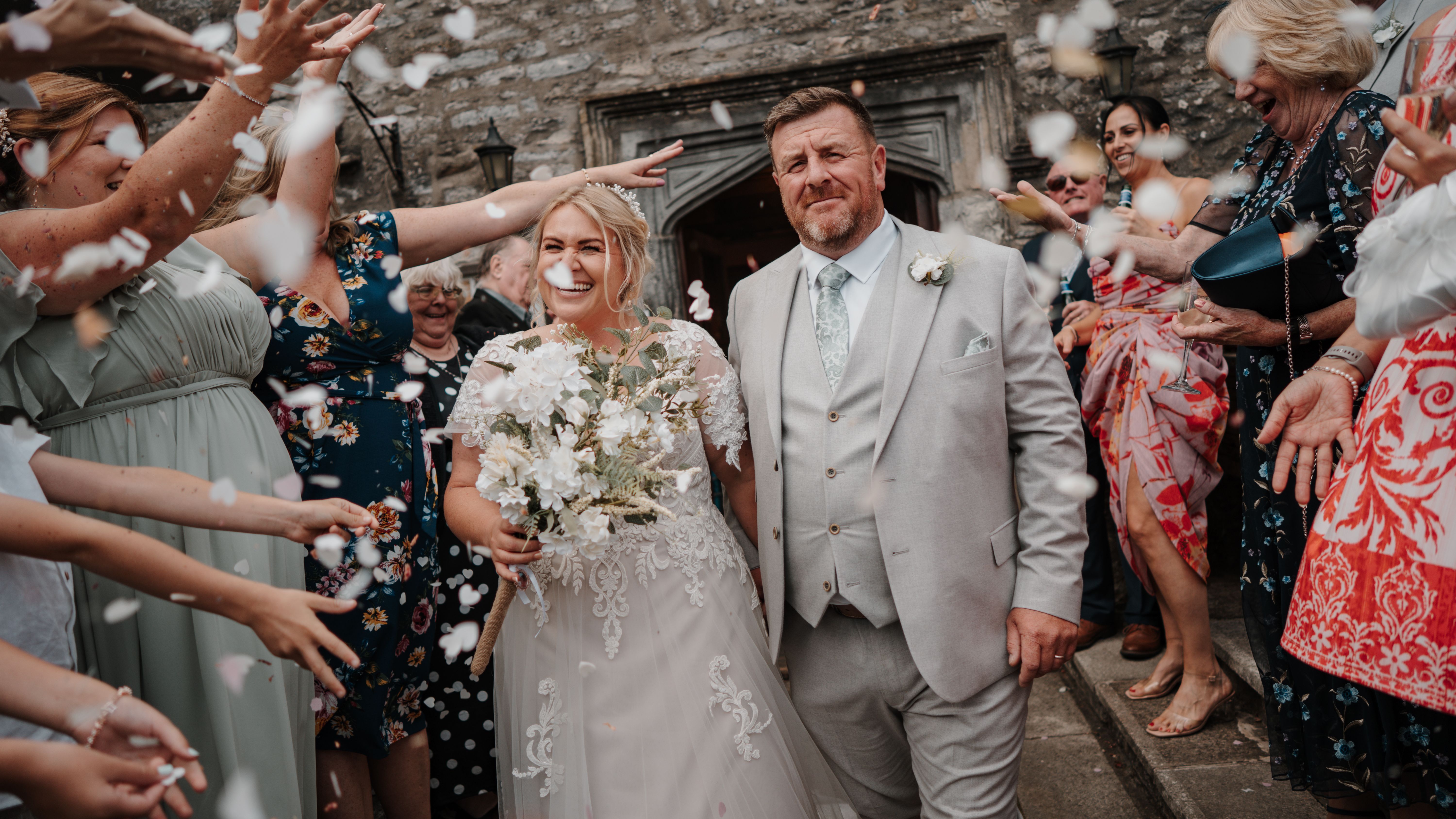 Bride in white lace wedding dress and groom in light grey suit walking out of a stone building, surrounded by guests throwing confetti.