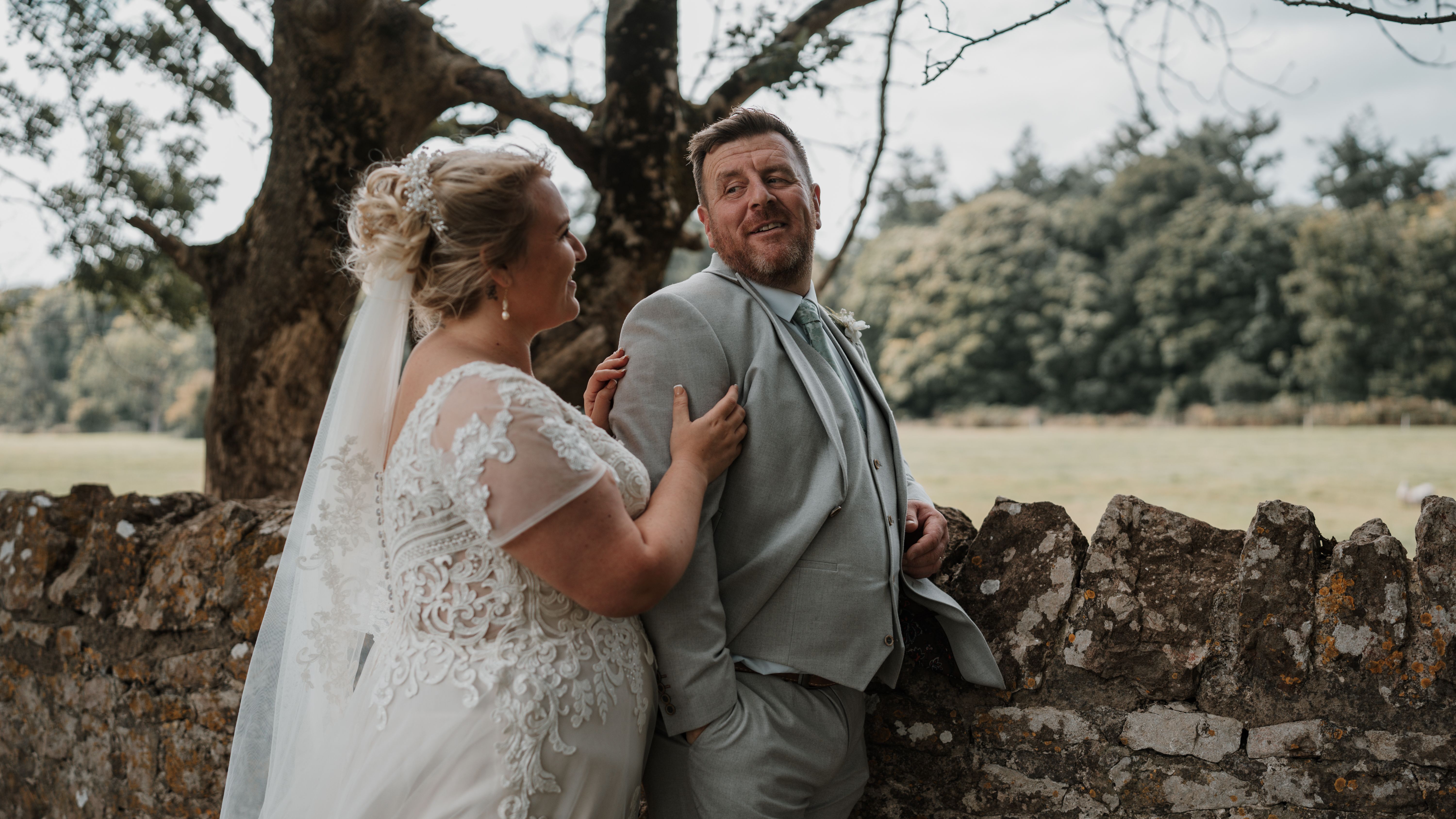 Bride and groom sharing a joyful moment outdoors, standing by a rustic stone wall with trees and greenery in the background.