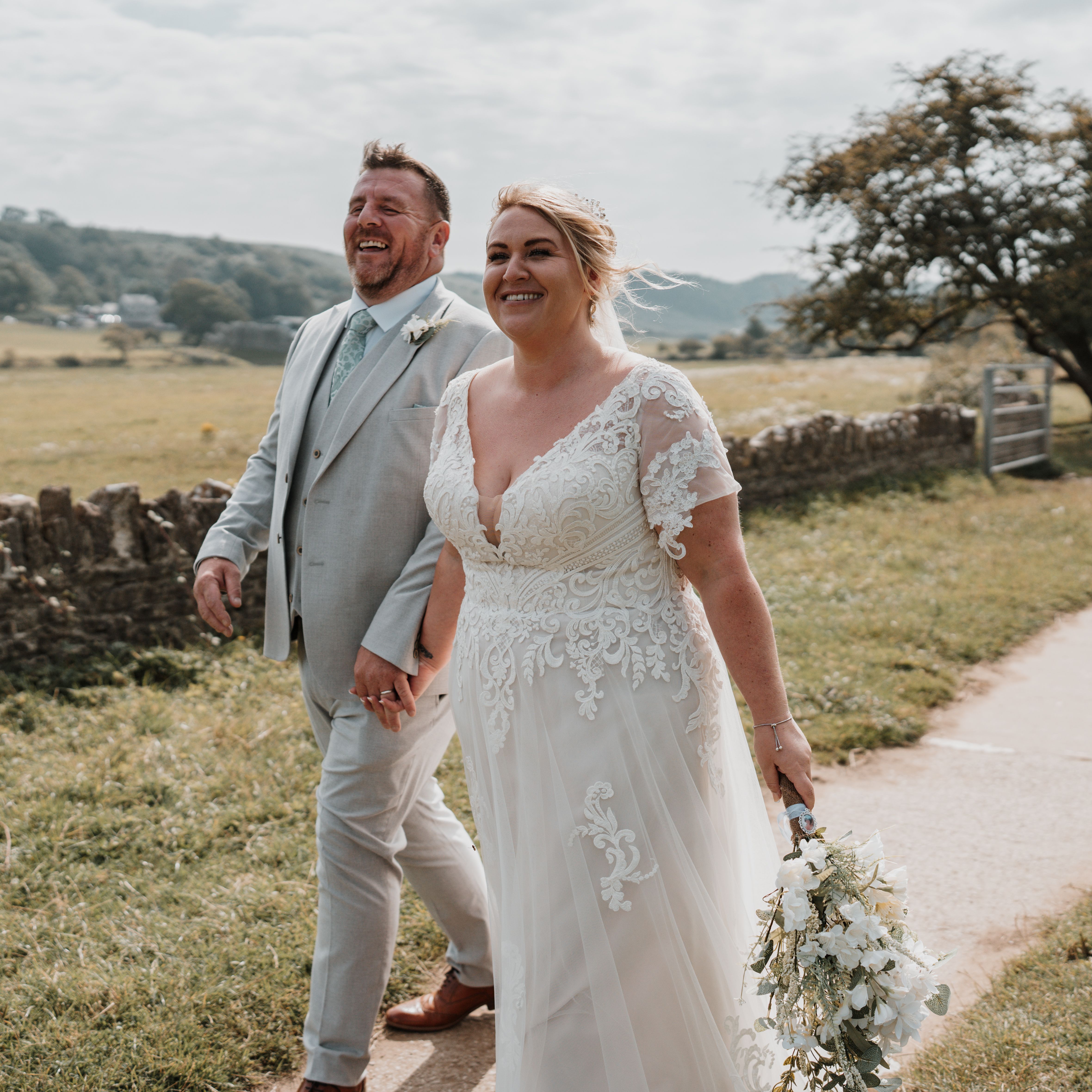 A bride and groom walk hand in hand outdoors, smiling on a sunny day with countryside scenery in the background.