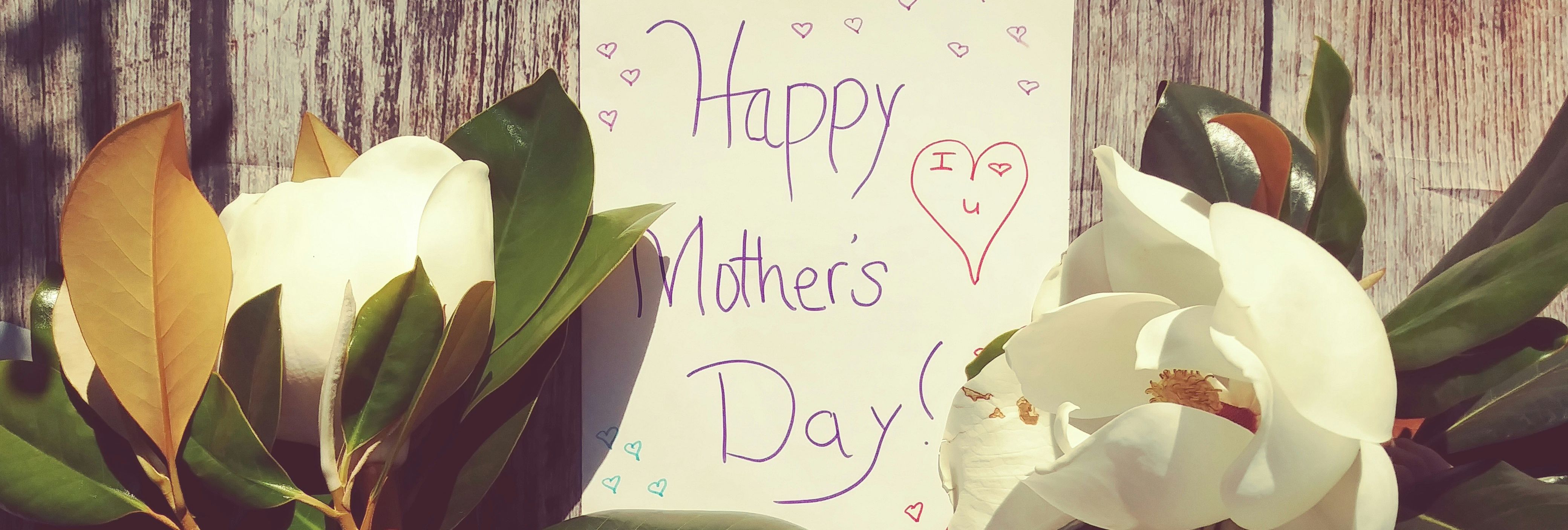 A child's hands hold large white magnolia flowers on either side of a handwritten 'Happy Mother's Day' card on a wooden background.