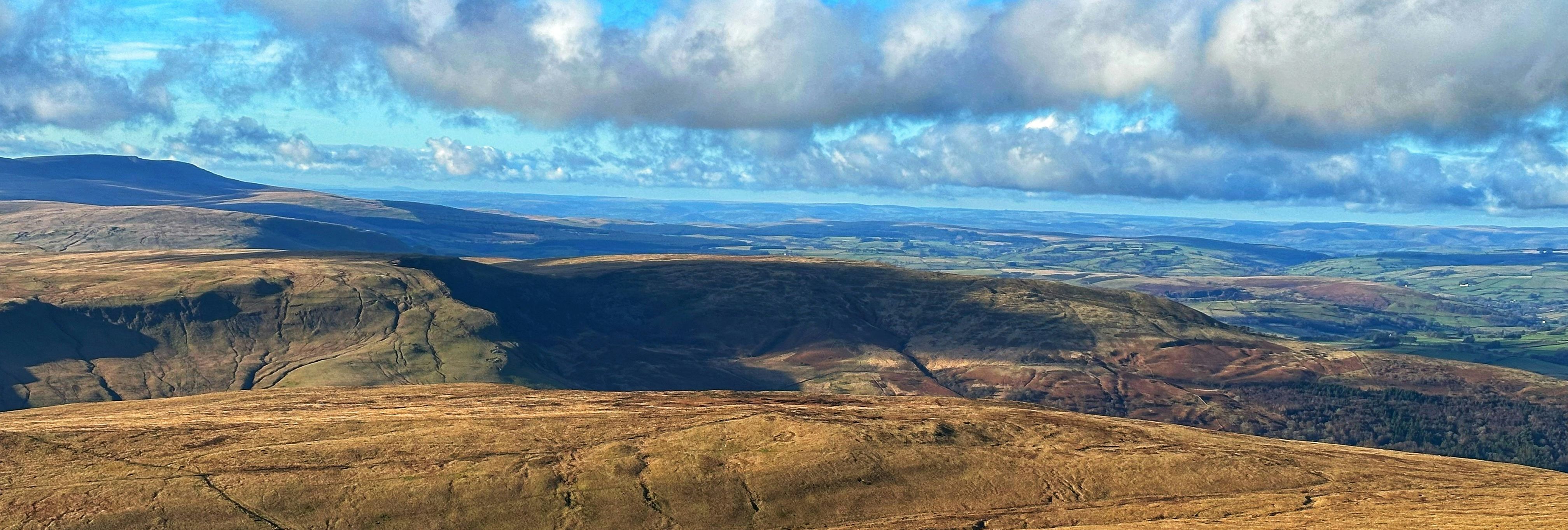 Two hikers walking across a vast grassy landscape with rolling hills under a blue sky with scattered clouds.