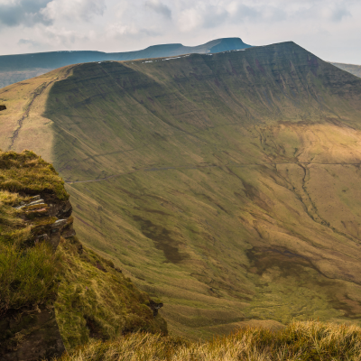 Grassy cliff edge overlooking a broad green valley and ridgeline under a cloudy sky