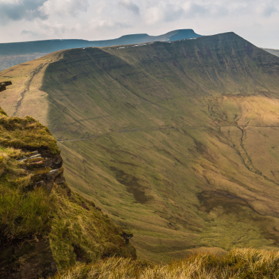 Grassy cliff edge overlooking a broad green valley and ridgeline under a cloudy sky