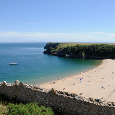 Sandy beach in a sheltered bay with a sailboat offshore and green cliffs under a clear blue sky.