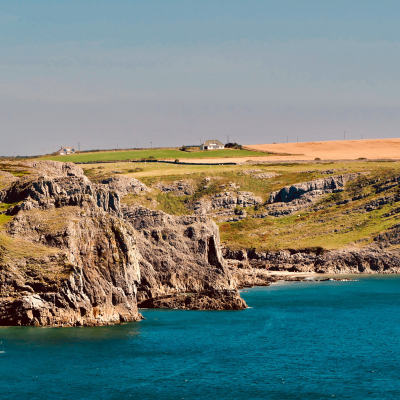 Rocky coastal cliffs beside turquoise sea with fields and a few buildings on a distant headland