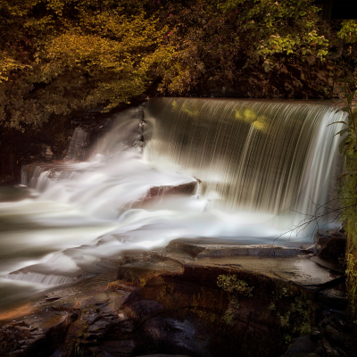 Long-exposure view of a small waterfall spilling over a stone weir into a rocky stream, framed by autumn trees.