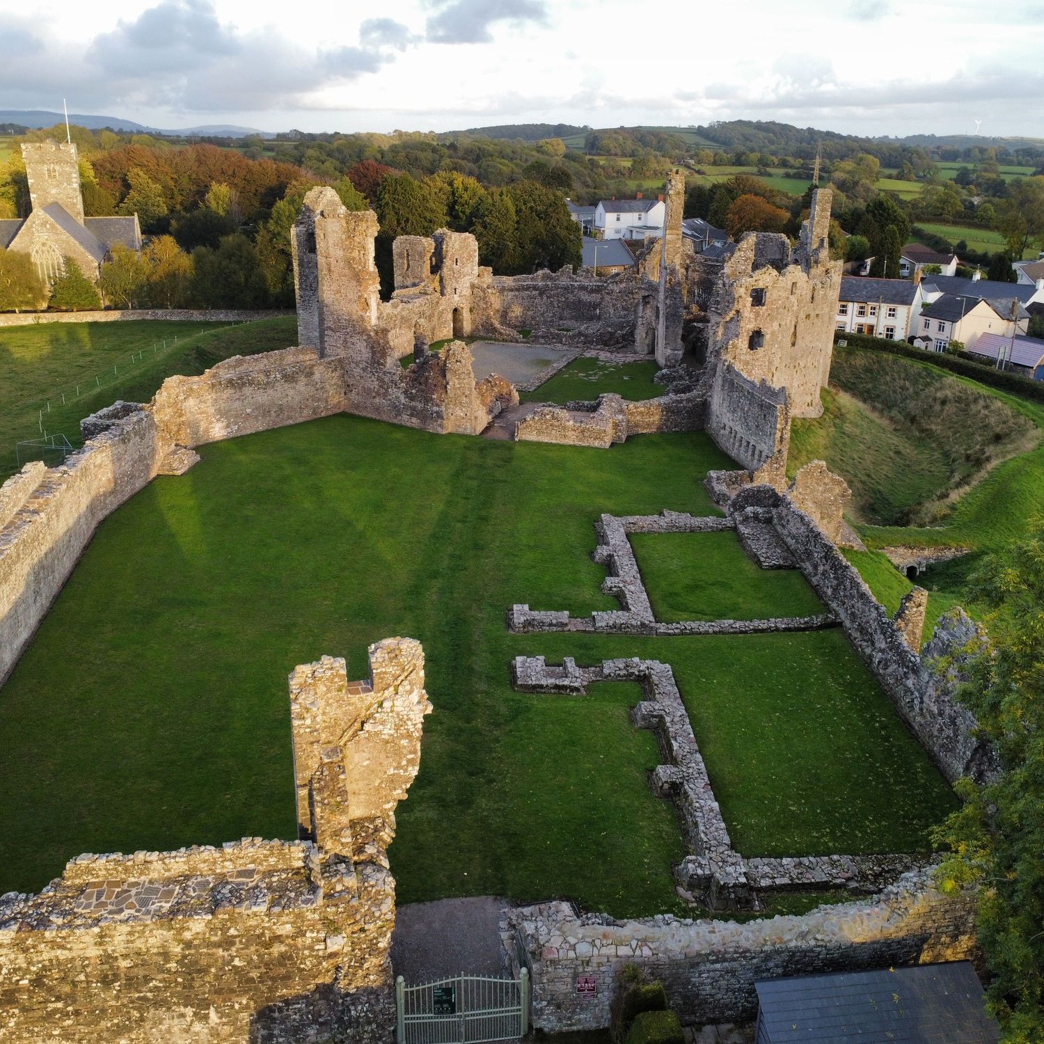 Aerial view of stone castle ruins with grassy courtyard beside a village and church, surrounded by rolling countryside.