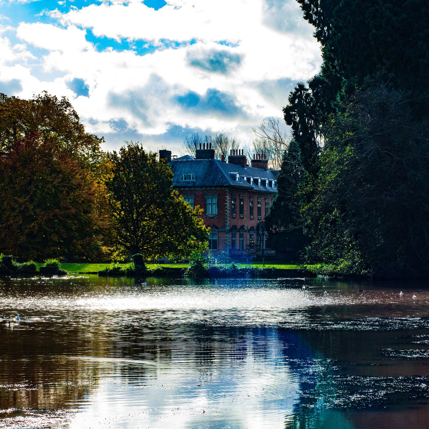 Historic brick manor house framed by autumn trees beside a lake under a cloudy sky