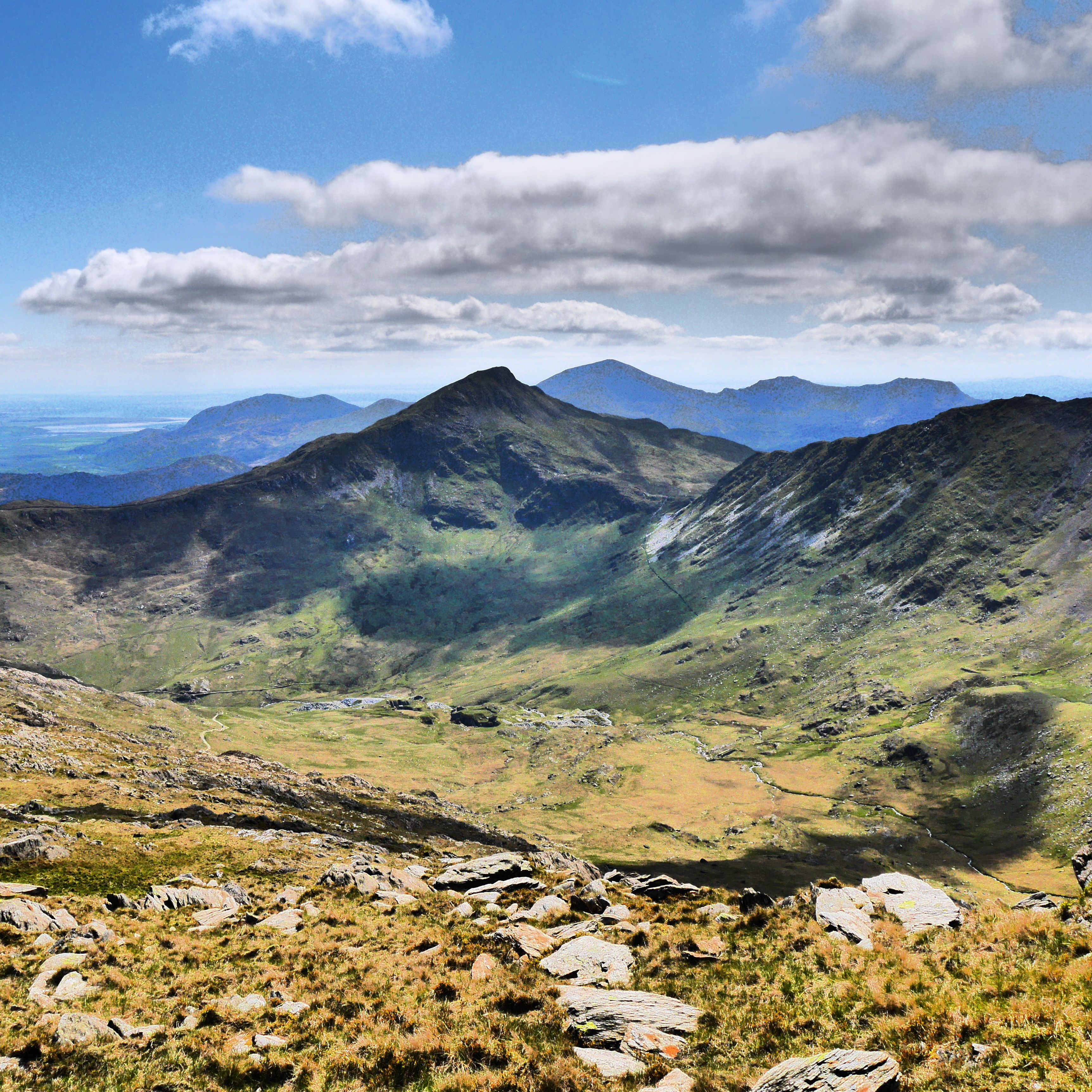 Mountain valley with rocky foreground and distant peaks under a blue sky with scattered clouds
