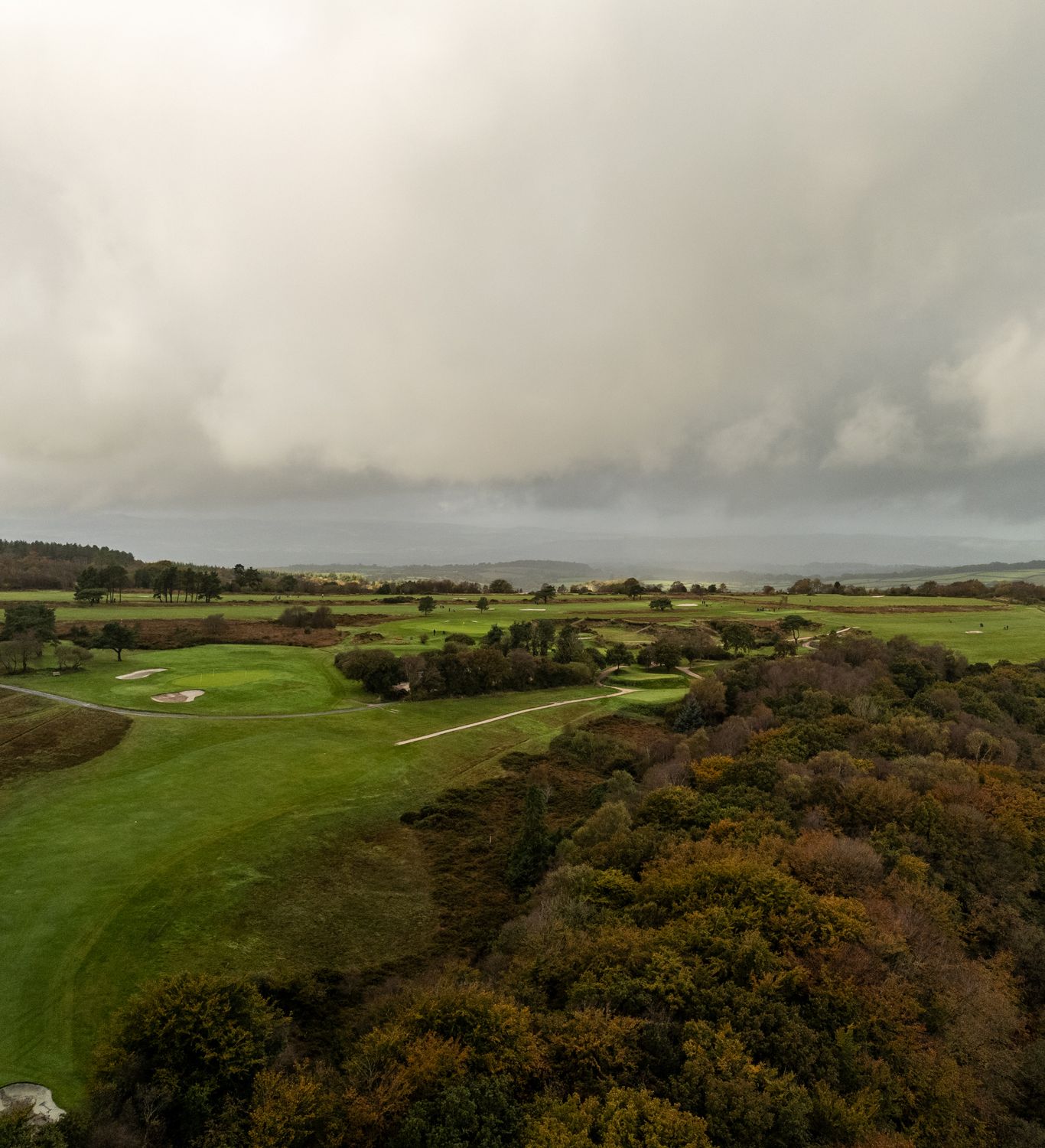 A scenic view of a golf course surrounded by rolling green fields, trees, and wooded areas, under a cloudy, overcast sky creating a serene and moody atmosphere.