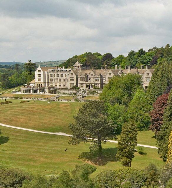 A grand countryside manor surrounded by lush green lawns, colorful gardens, and dense trees, set against rolling hills and a partly cloudy sky.