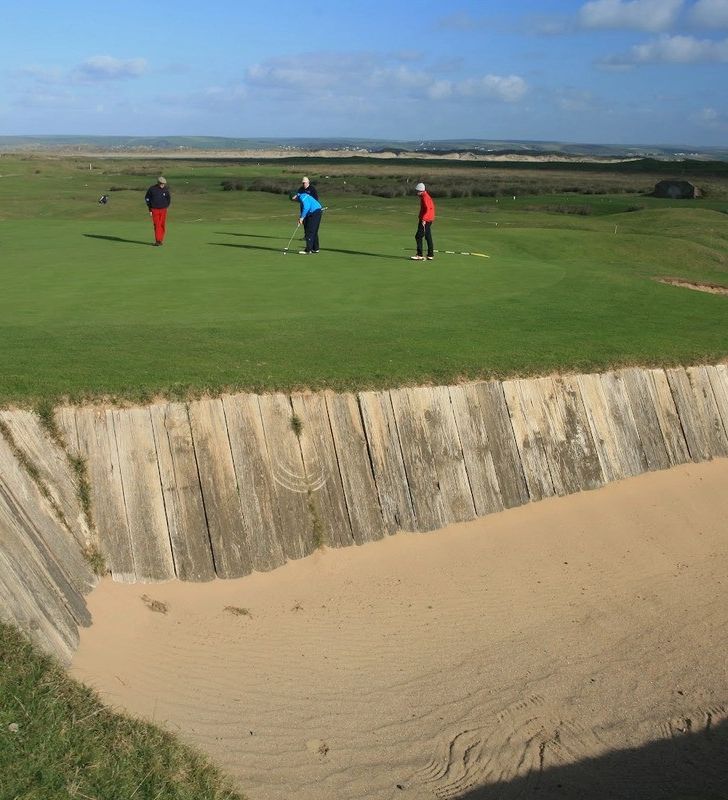 A grassy golf course featuring a deep sand bunker reinforced with wooden planks, with three golfers standing on the green under a sunny sky with scattered clouds.