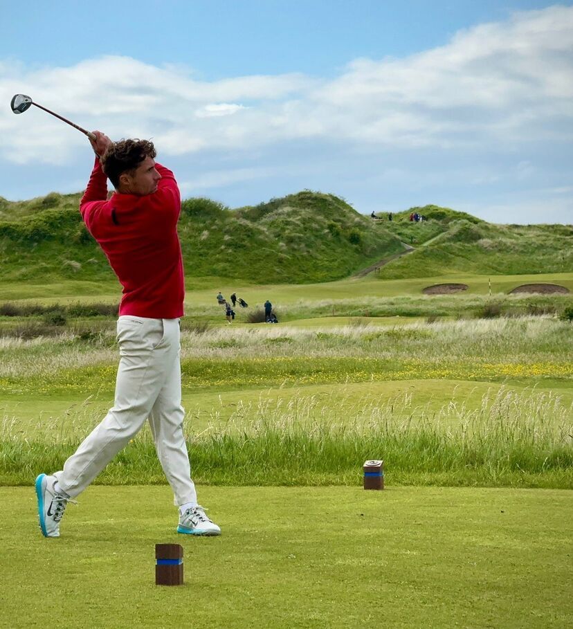 A golfer in a red sweater and white pants completes a powerful swing on a grassy tee box, set against rolling dunes and a bright, partly cloudy sky.